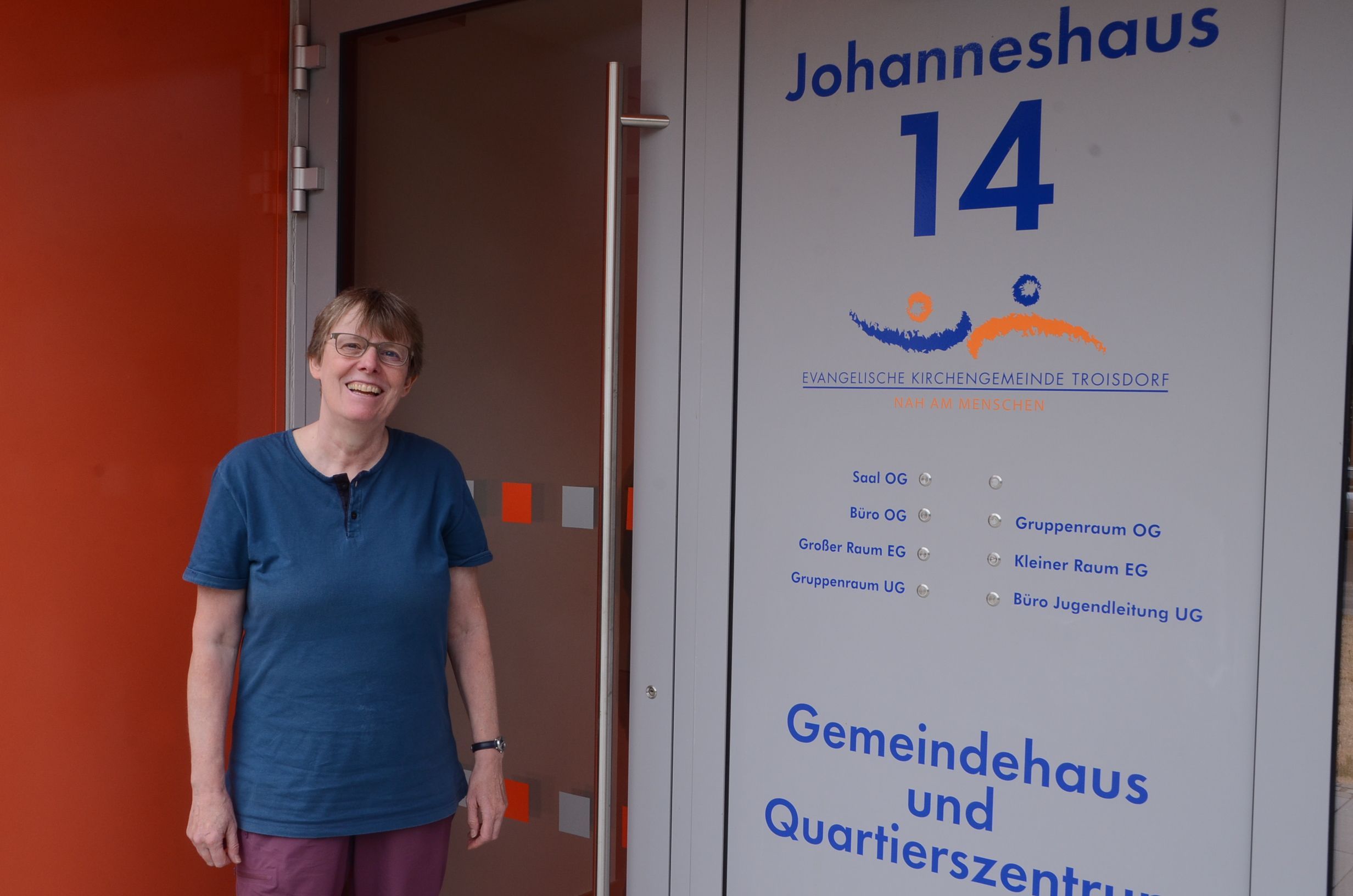 Eine Frau steht vor deim Eingang in ein Gemeindehaus; es gibt Klingeln für viele Räume.