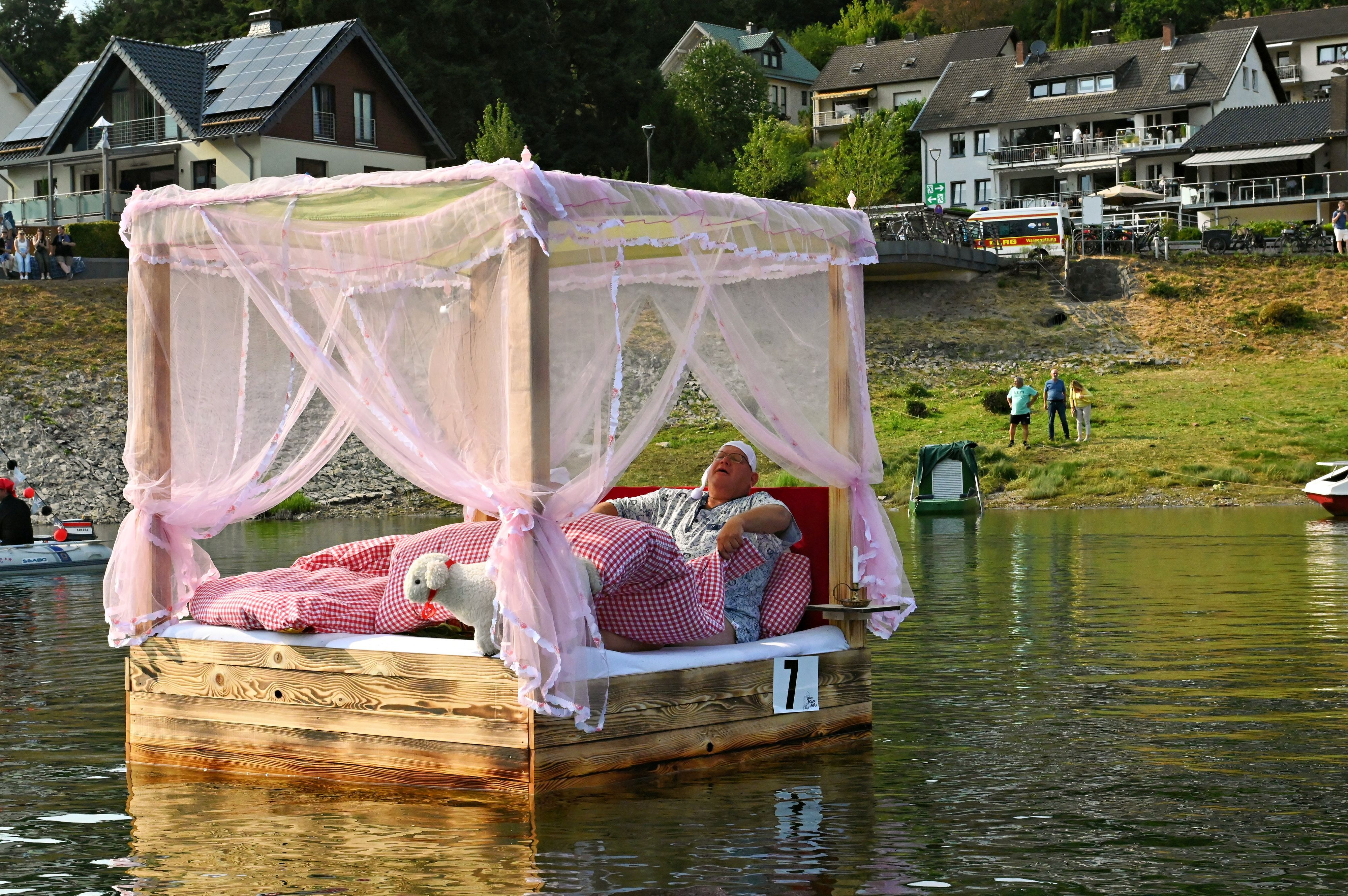 Auf dem Wasser dümpelt ein Himmelbett mit rotkariertem Bettzeug, in dem ein Mann im Schlafanzug liegt.