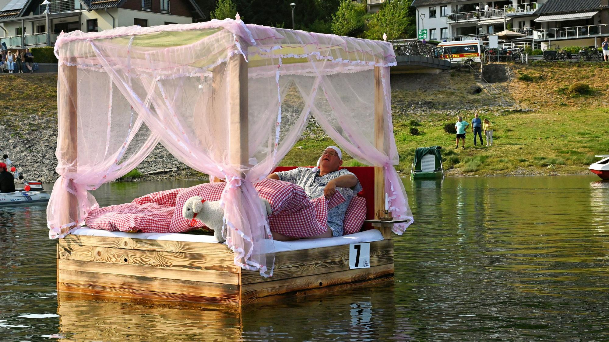 Auf dem Wasser dümpelt ein Himmelbett mit rotkariertem Bettzeug, in dem ein Mann im Schlafanzug liegt.