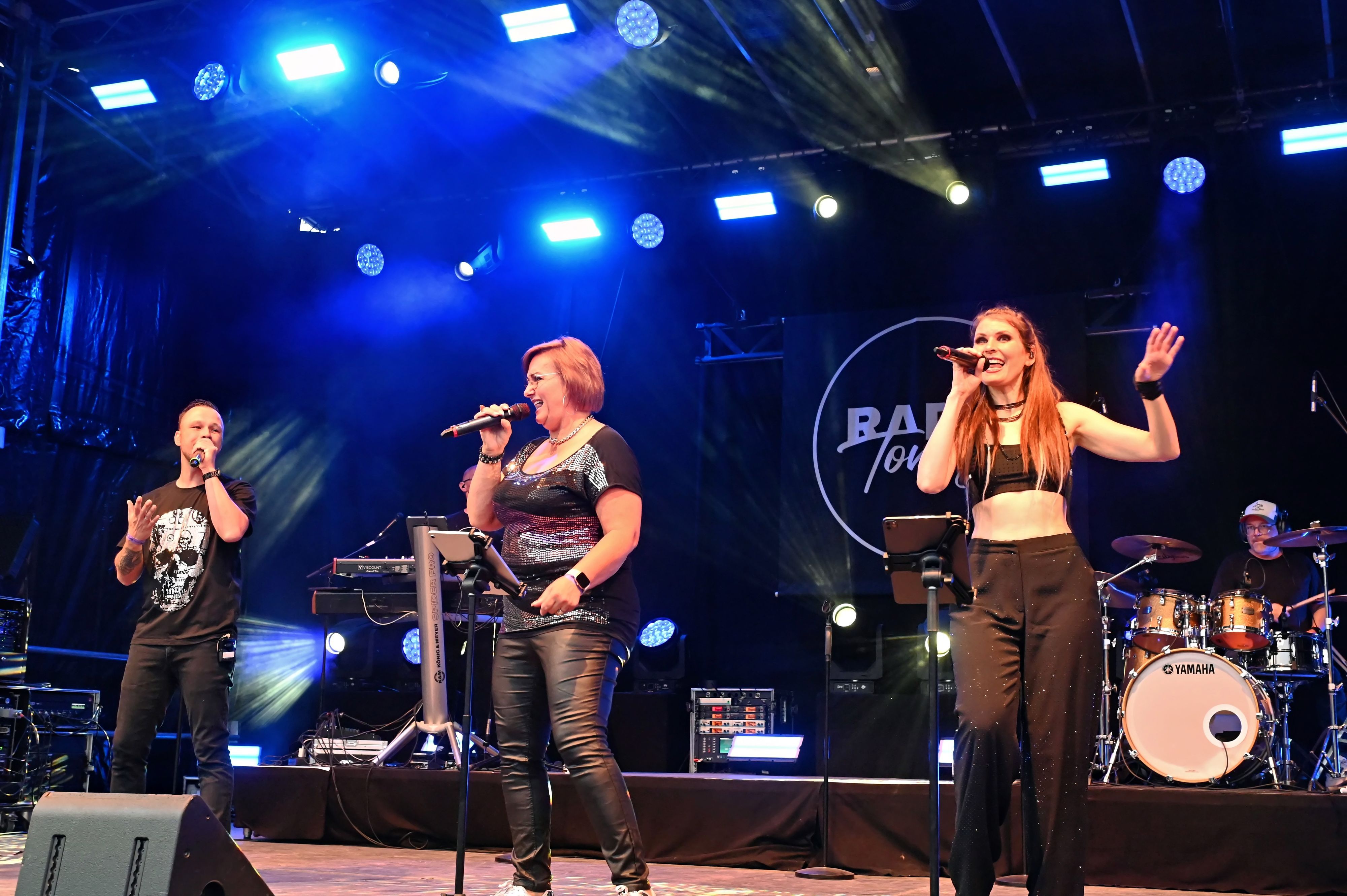Auf der Bühne stehen Mitglieder der Band, im Vordergrund zwei Sängerinnen.