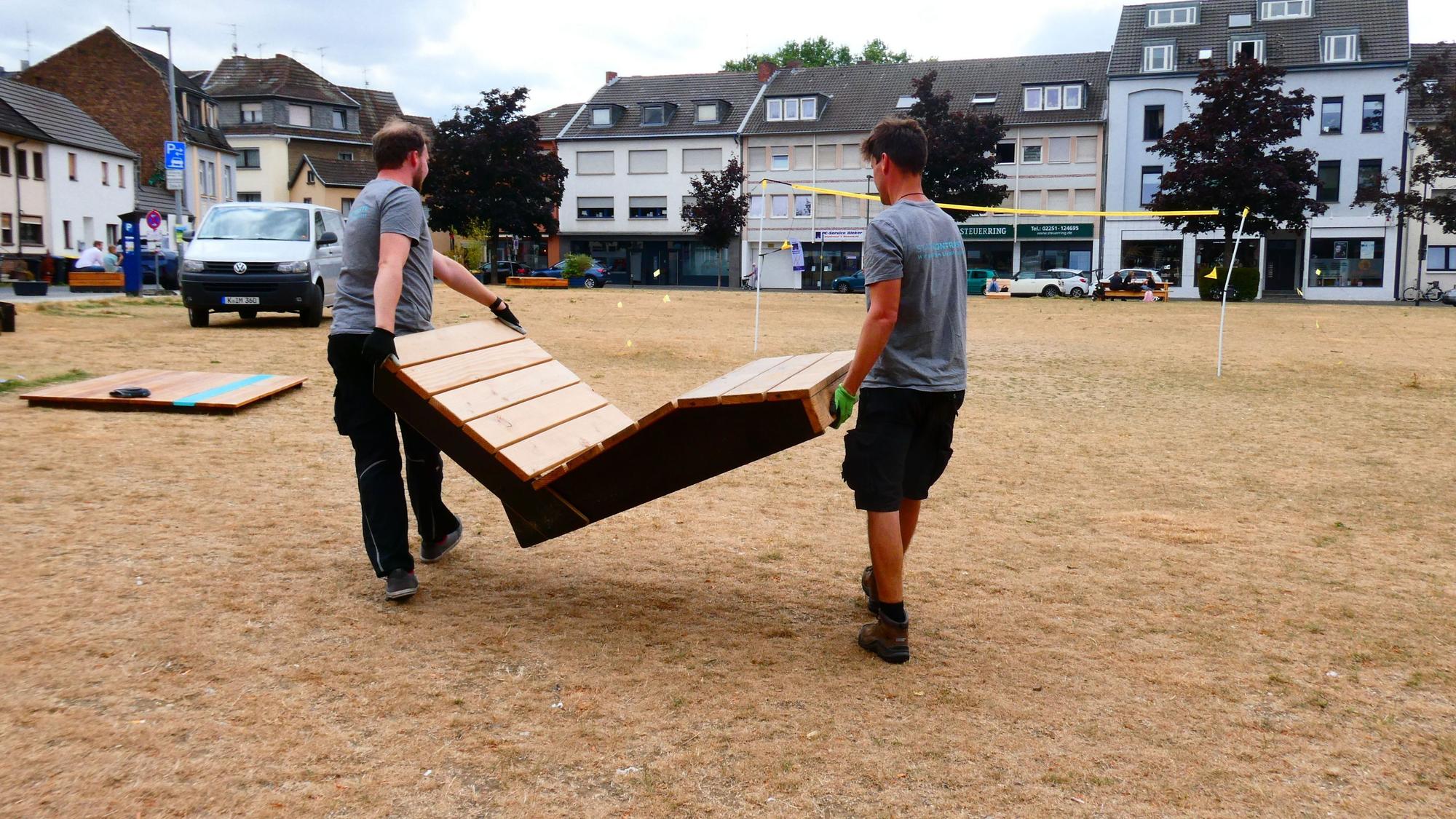Das Bild zeigt zwei Männer, die eine Holzbank wegtragen.