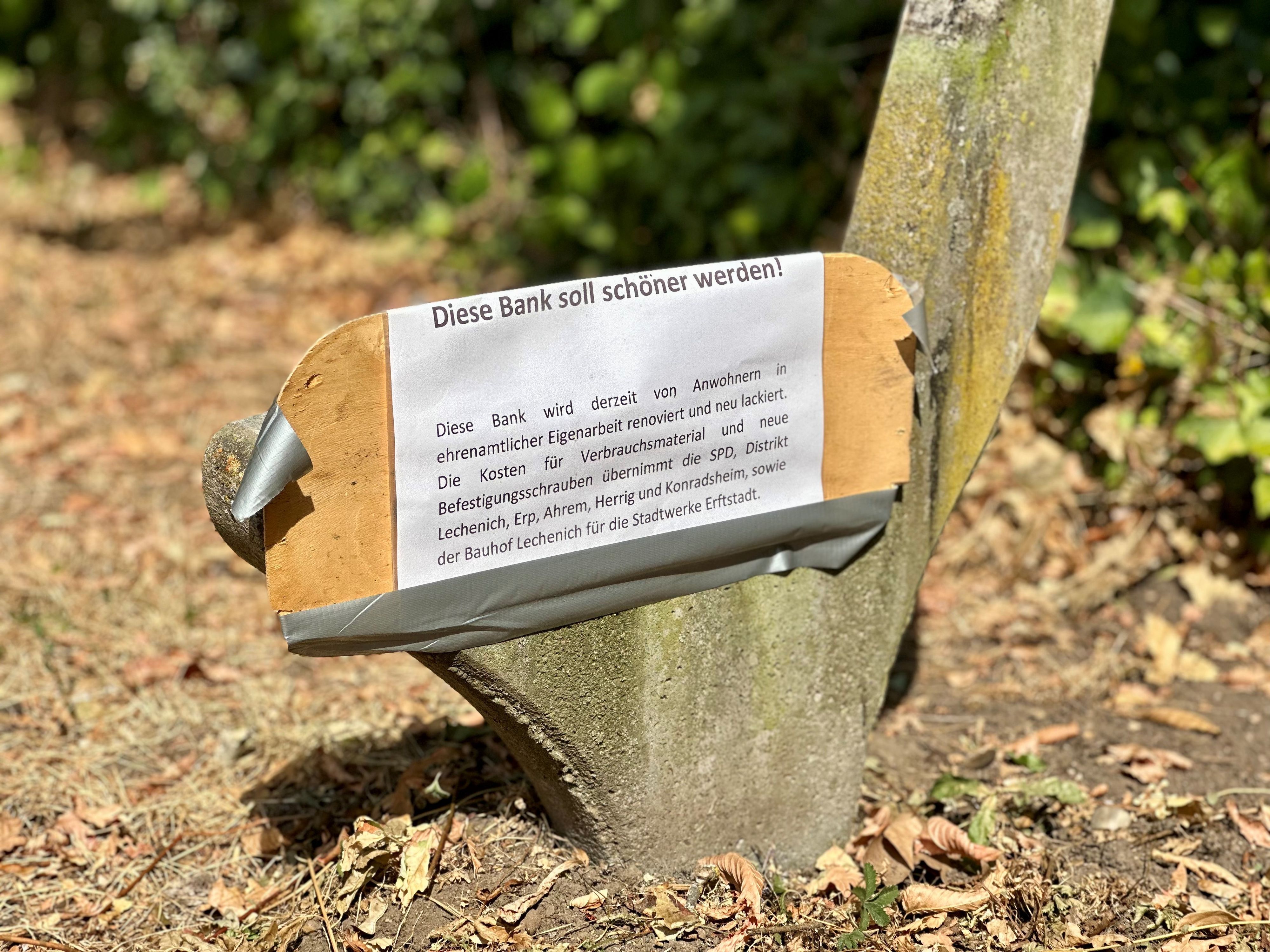 Am Betonfundament einer Parkbank hängt ein weißes Schild.