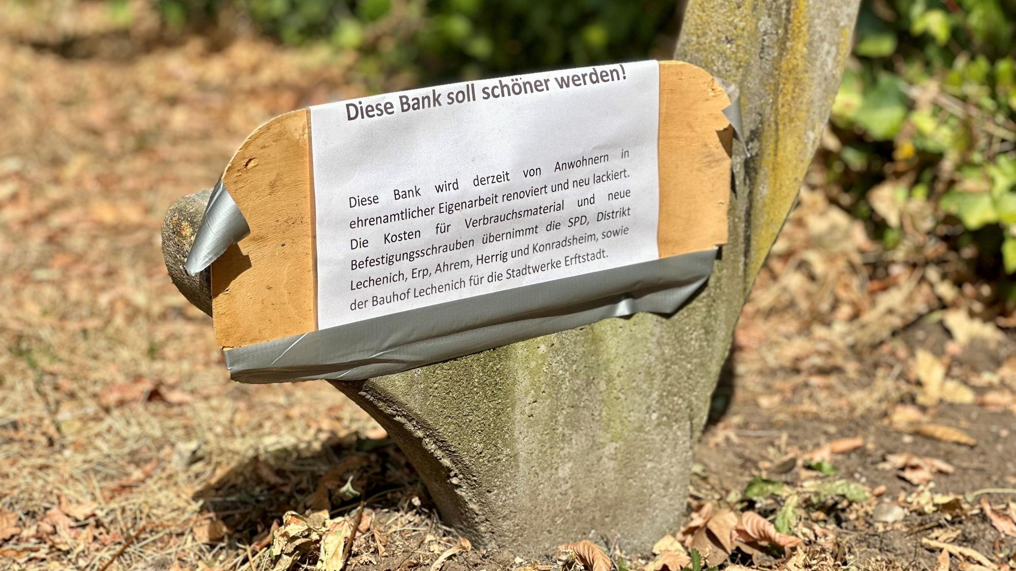 Am Betonfundament einer Parkbank hängt ein weißes Schild.