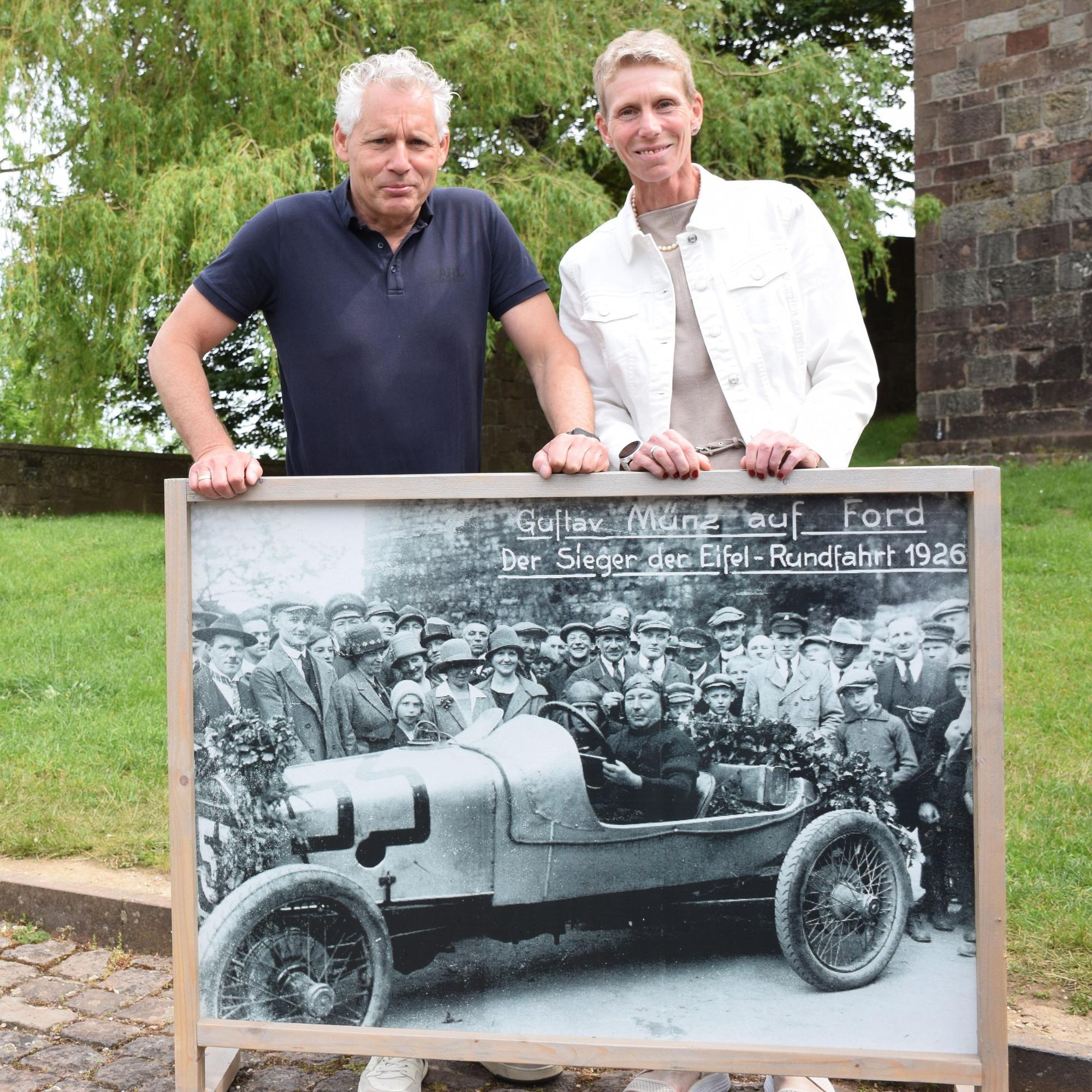 Georg Münz und Jette Bork-Wagenblast halten ein großformatiges Foto, das Gustav Münz als Sieger der Eifel-Rundfahrt 1926 in seinem Ford zeigt.