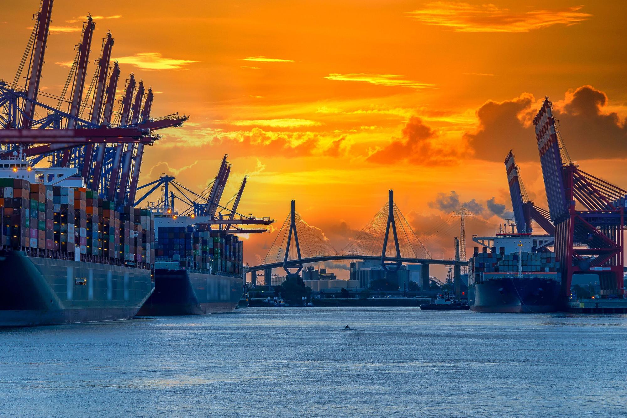 Containerschiffe, Hafenkräne und eine Brücke in der Ferne. Orangefarbener Morgenhimmel im Hintergrund.