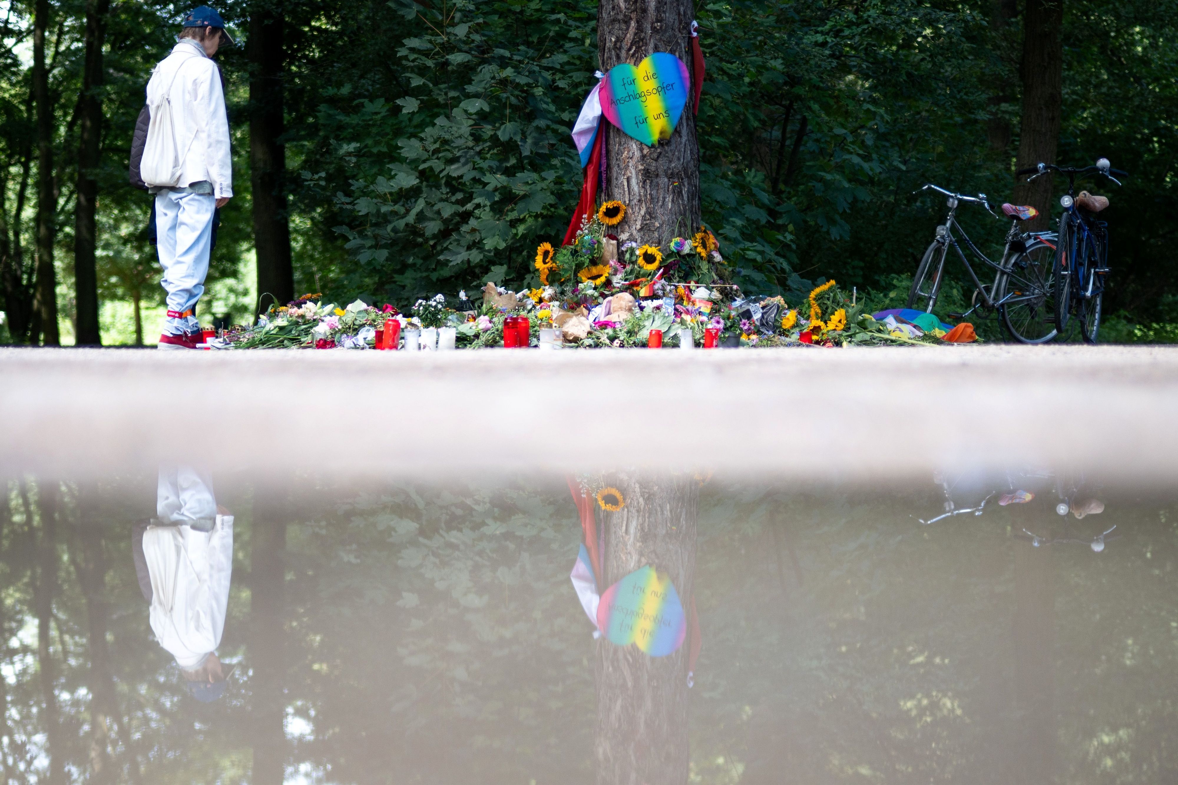 Trauernde stehen im Berliner Tiergarten an einem Baum, an dem unterschiedliche Gegenstände zum Gedenken an die Opfer des Anschlags auf den CSD abgelegt wurden.