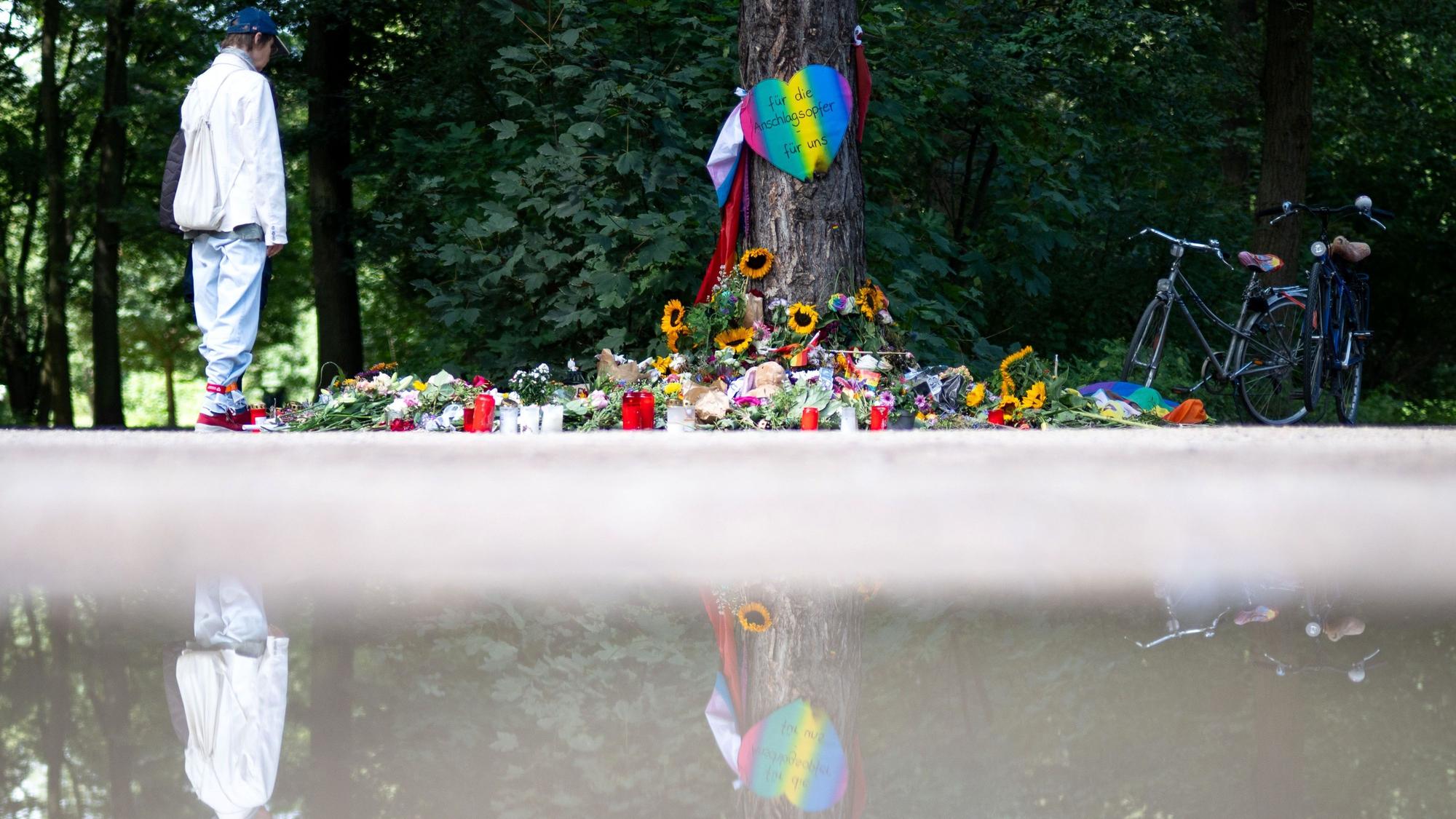 Trauernde stehen im Berliner Tiergarten an einem Baum, an dem unterschiedliche Gegenstände zum Gedenken an die Opfer des Anschlags auf den CSD abgelegt wurden.