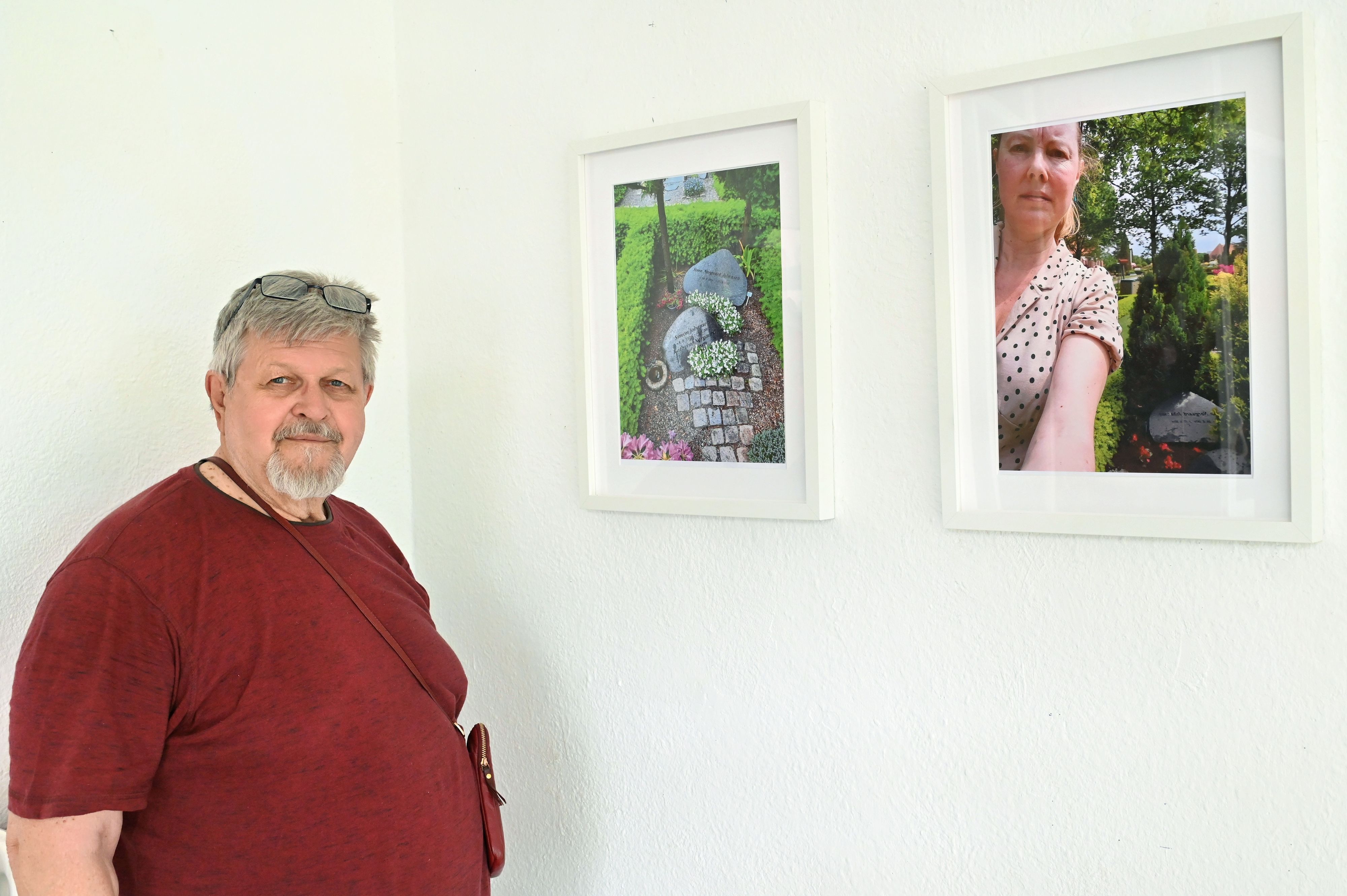 Ein bärtiger Mann in rotem T-Shirt steht neben zwei Fotografien, die gerahmt an der Wand hängen.
