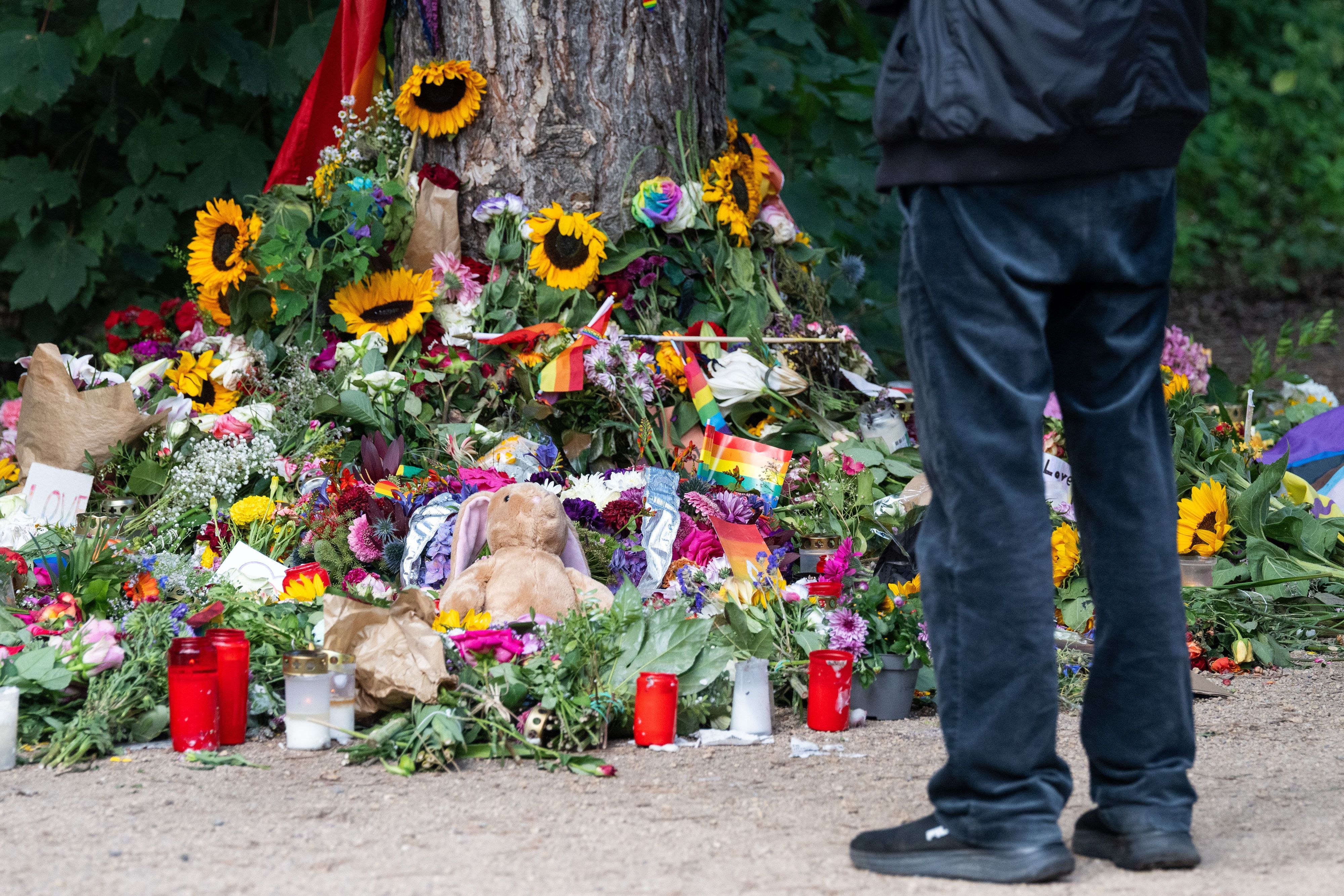 Trauernde stehen im Berliner Tiergarten an einem Baum, an dem unterschiedliche Gegenstände zum Gedenken an die Opfer des Anschlags auf den CSD abgelegt wurden.