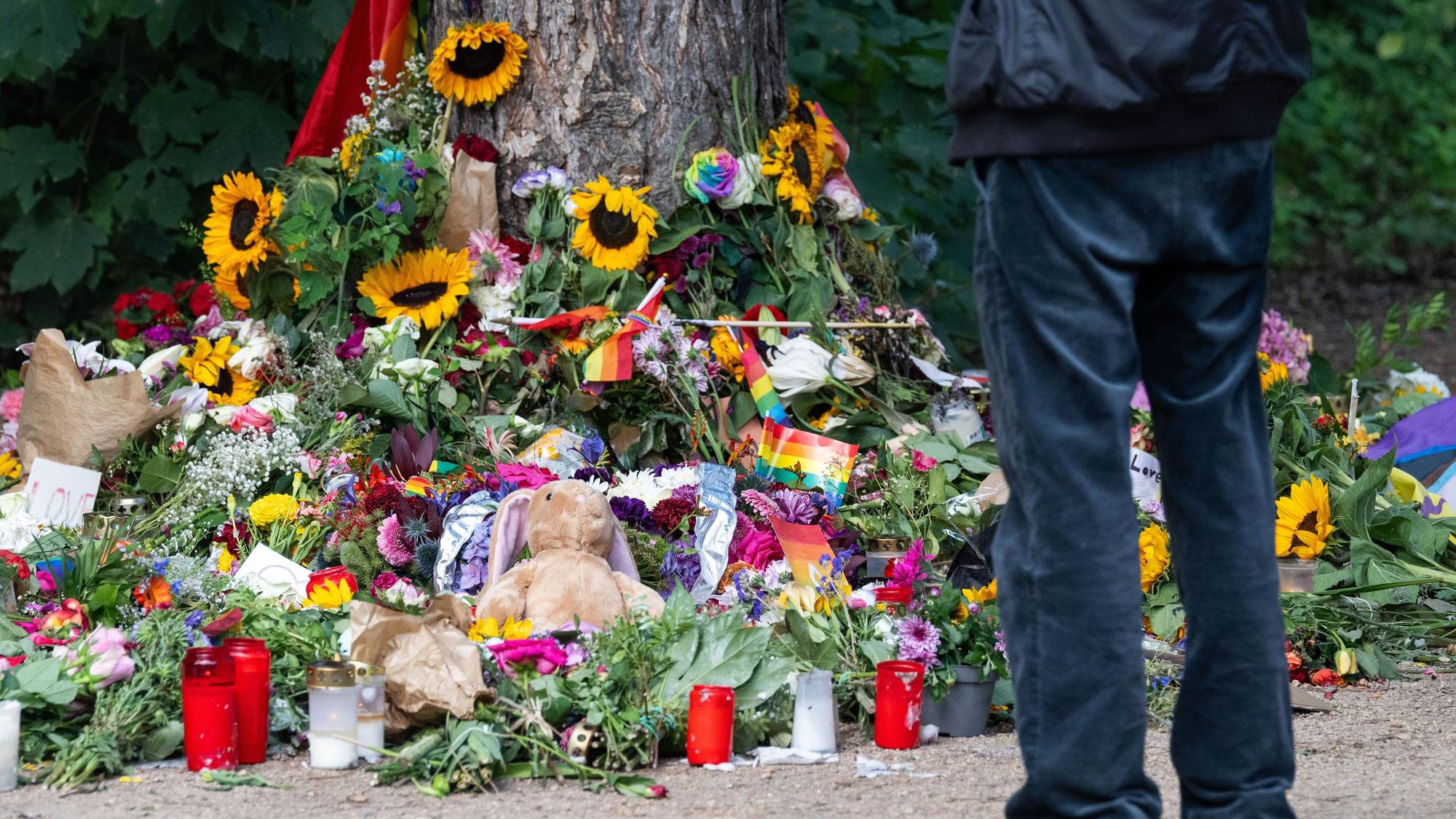 Trauernde stehen im Berliner Tiergarten an einem Baum, an dem unterschiedliche Gegenstände zum Gedenken an die Opfer des Anschlags auf den CSD abgelegt wurden.