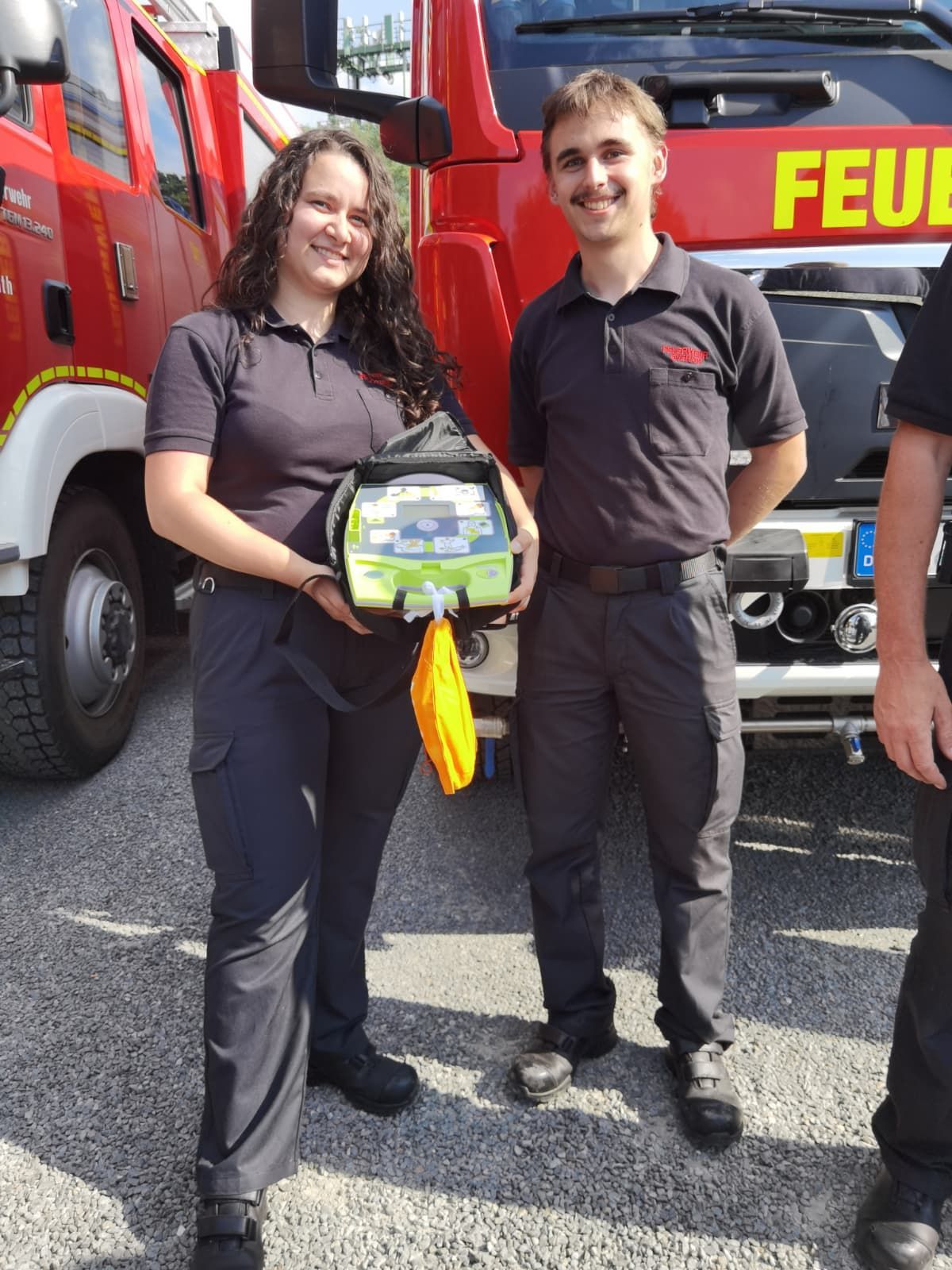 Zu sehen sind eine Feuerwehrfrau und ein Feuerwehrmann nebeneinander und sie lächeln. Sie hält einen Defibrillator in der Hand.