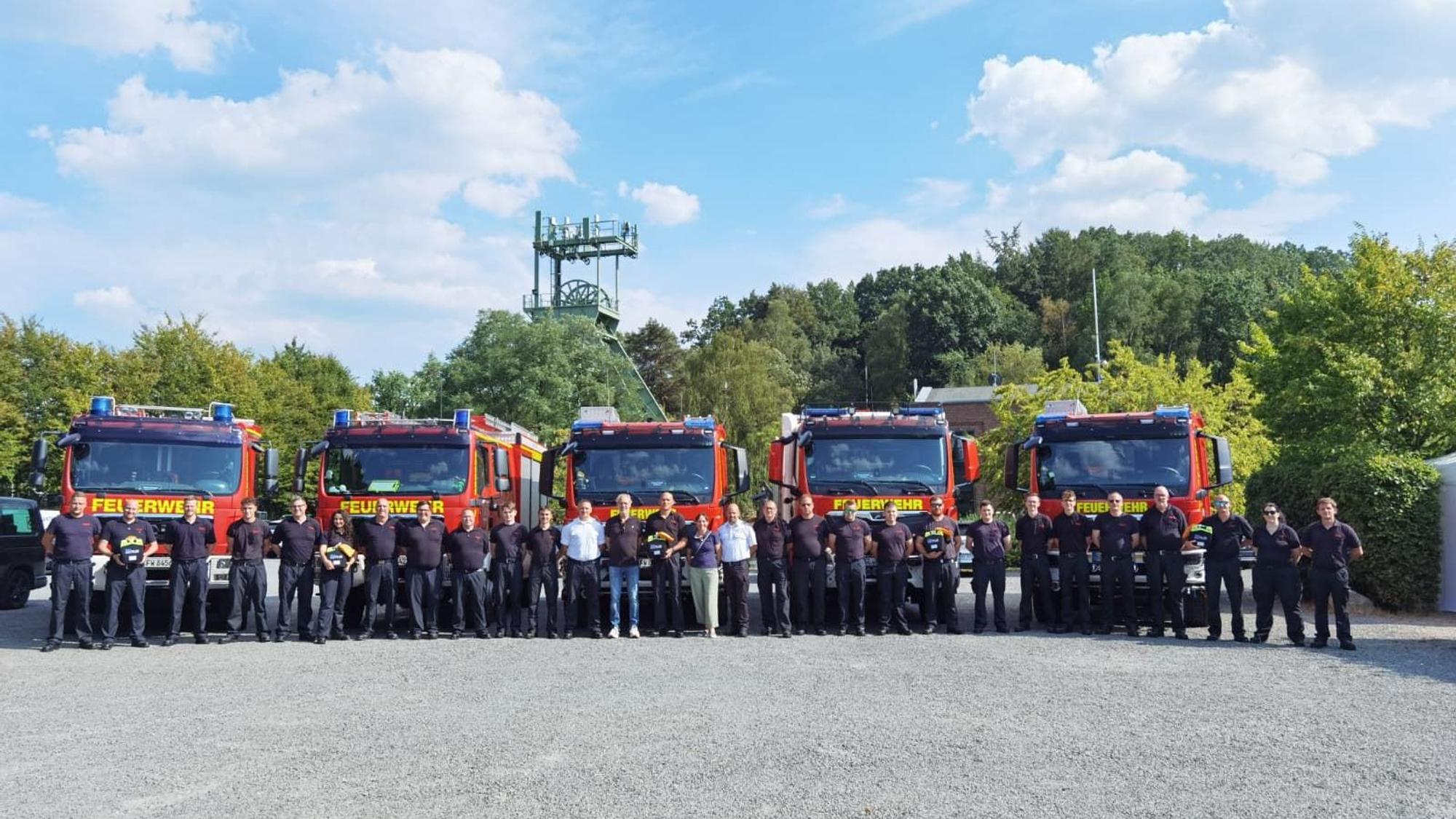 Zu sehen sind viele Feuerwehrleute aufgereiht nebeneinander. Hinter ihnen stehen fünf Feuerwehrwagen.