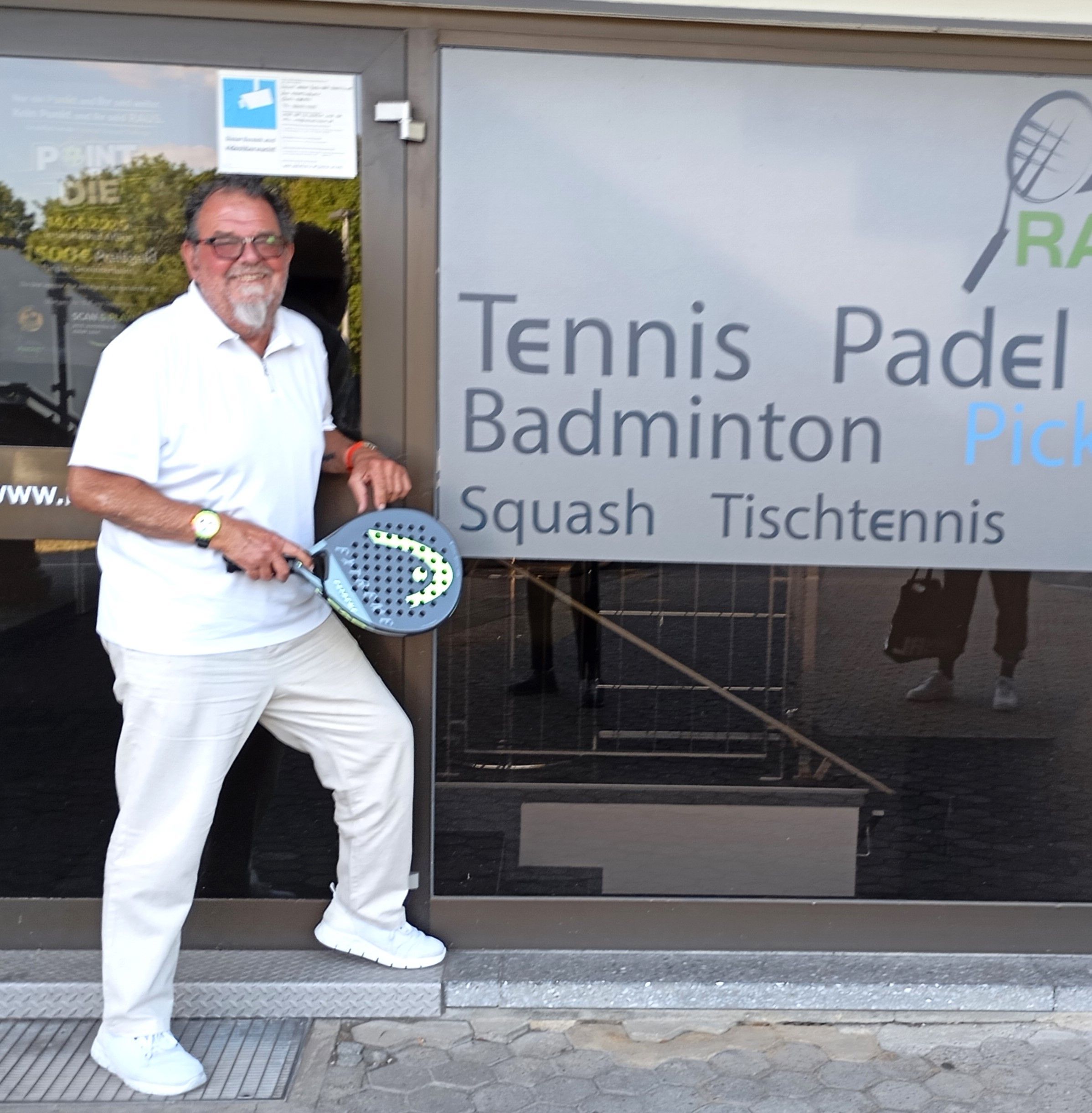Das Foto zeigt einen Mann mit einem Tennisschläger vor einer Tennis- und Badminton-Halle.