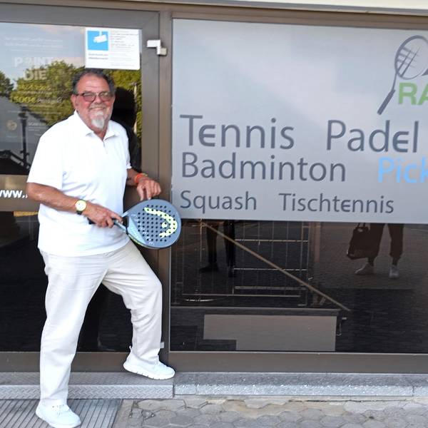 Das Foto zeigt einen Mann mit einem Tennisschläger vor einer Tennis- und Badminton-Halle.