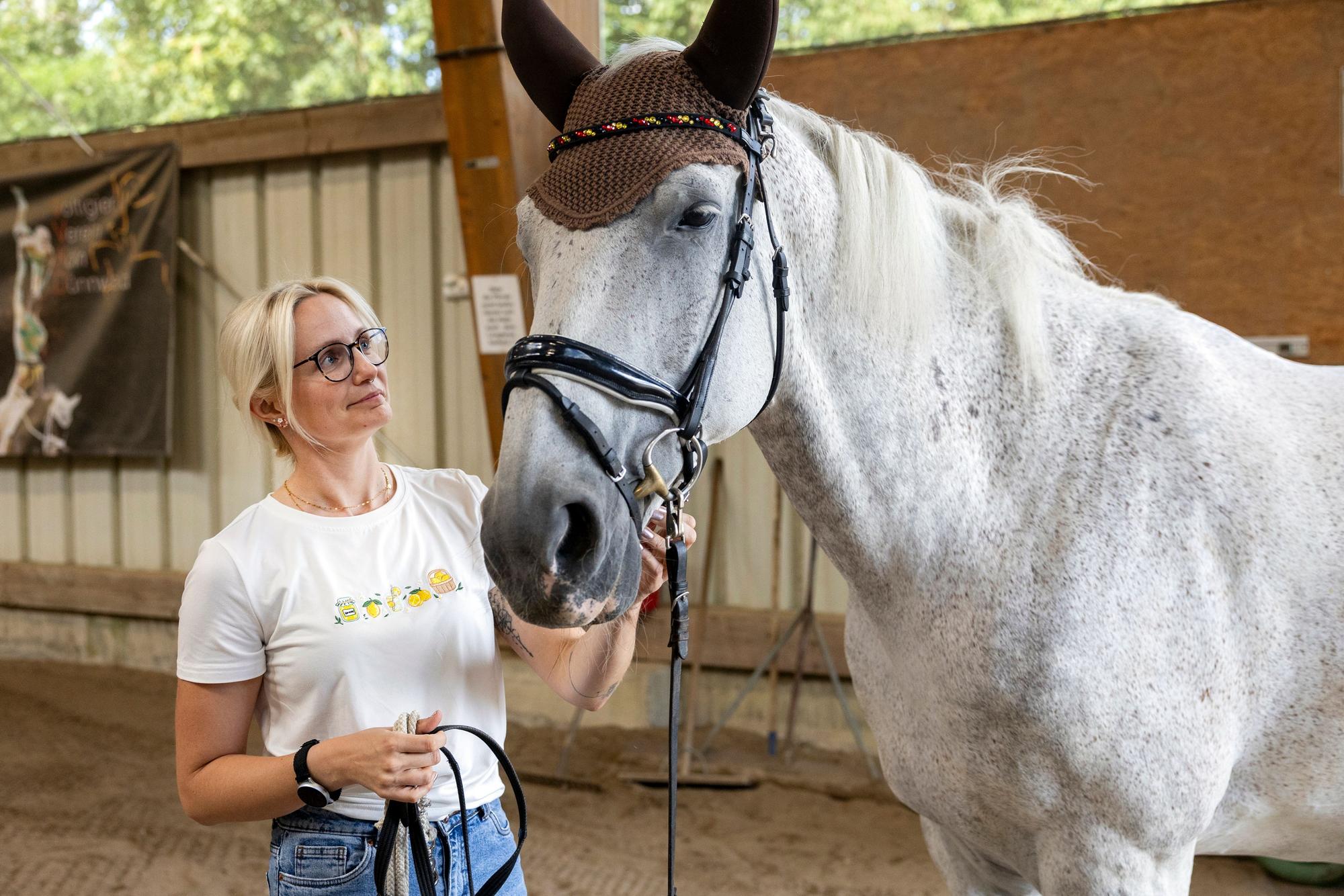 Eine Frau mit blondem Haar und Brille steht in Jeans und T-Shirt neben einem Schimmel.