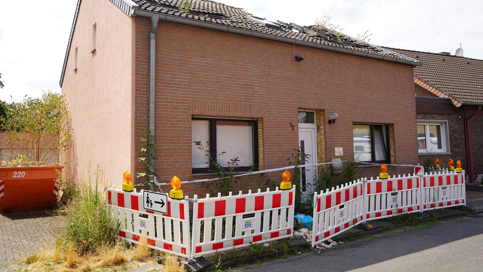 Das Bild zeigt ein Haus mit abgebranntem Dachstuhl. Davor ist der Gehweg abgesperrt.