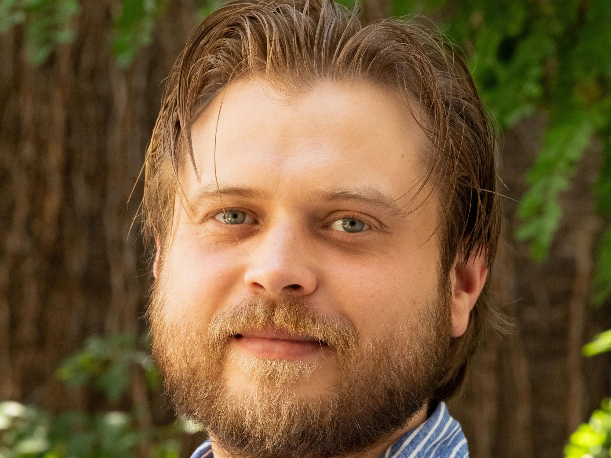 Ein Porträt von Dominik Zell im Freien. Er trägt einen Bart und ein blau-weiß-gestreiftes Hemd.
