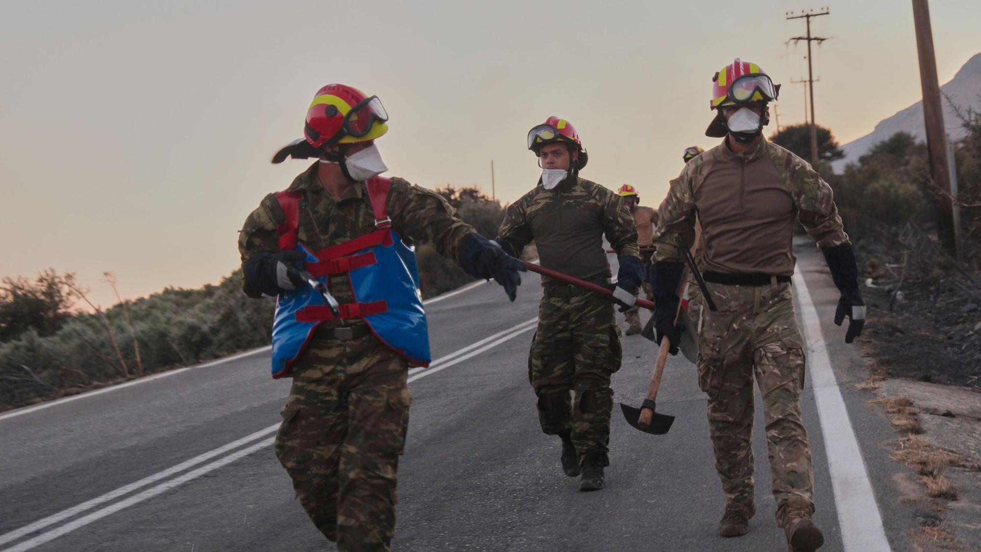 Feuerwehrleute gehen während eines Waldbrands in der Region Rethymno auf der südgriechischen Insel Kreta mit Werkzeugen eine Straße entlang.