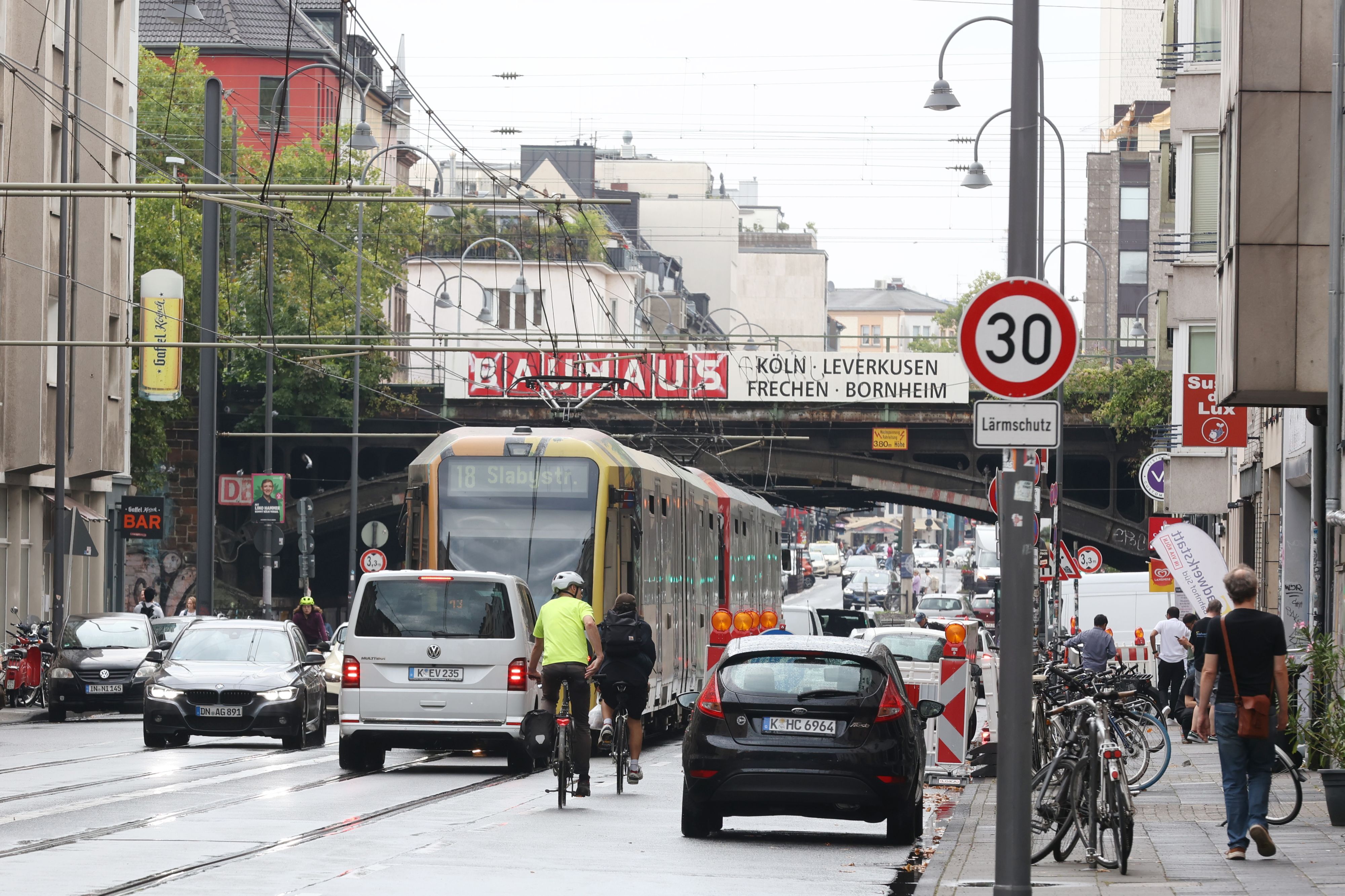 Das Foto zeigt die Luxemburger Straße auf Höhe der Eisenbahnbrücke Köln Süd. Die Straße wird genutzt von Straßenbahnen, Autos, Radfahrern und Fußgängern.