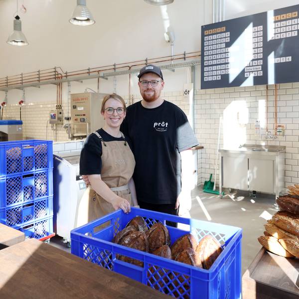 Eine Frau mit Zopf, Brille und Schürze steht neben einem Mann mit Cap und Bart. Sie stehen in einer großen Halle hinter einer Verkaufstheke, vor und neben sich sind Kisten mit Broten,