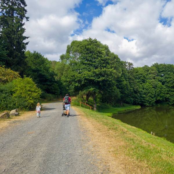 Ein Mann schiebt einen Kinderwagen über einen Weg neben einem See, links von ihm läuft ein Mädchen.