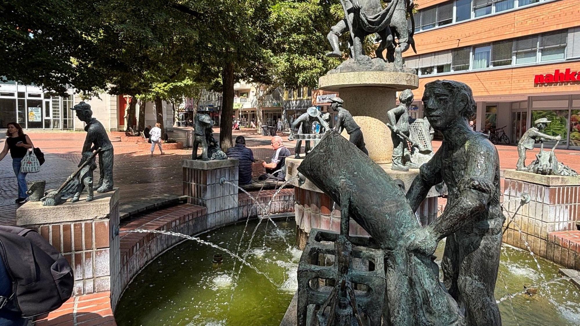 Das Bild zeigt einen Brunnen mit Figuren auf einem Platz.
