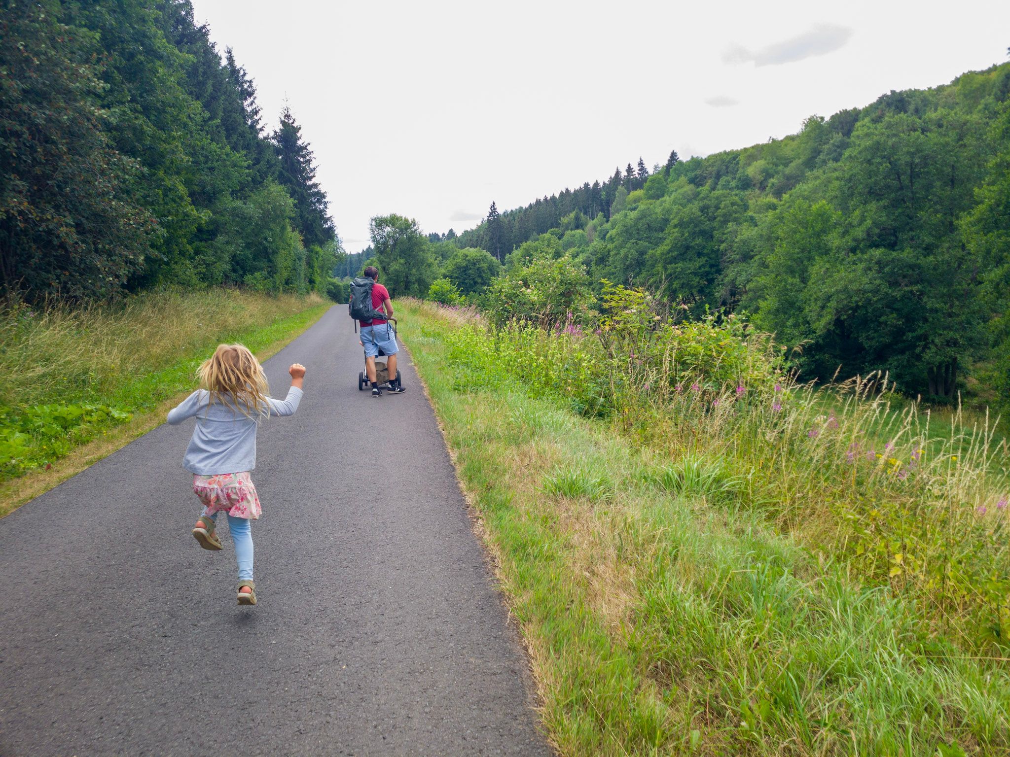 Ein Mädchen hüpft über einen Weg, vor ihr läuft ein Mann mit Kinderwagen.