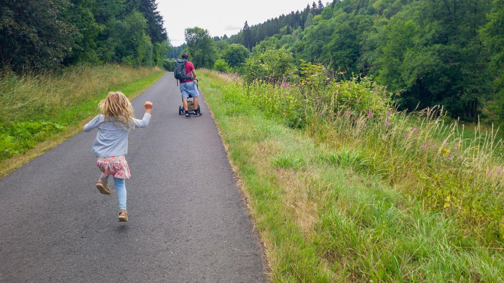 Ein Mädchen hüpft über einen Weg, vor ihr läuft ein Mann mit Kinderwagen.