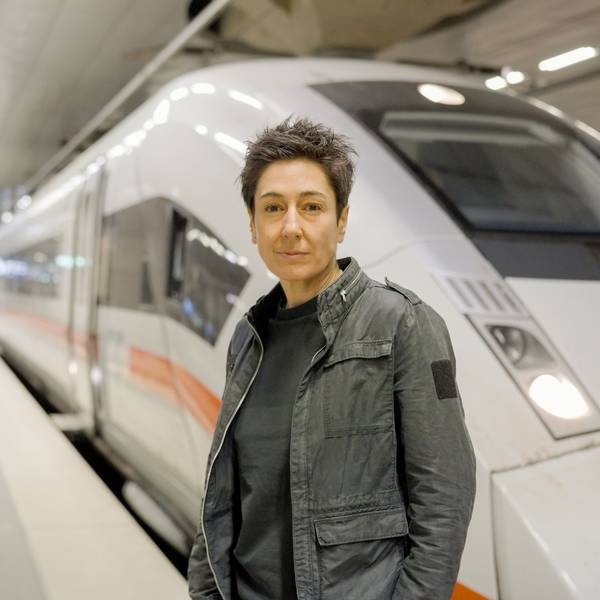 Dunja Hayali steht in einer Bahnhofshalle auf einem leeren Gleis vor einem ICE und schaut in die Kamera.
