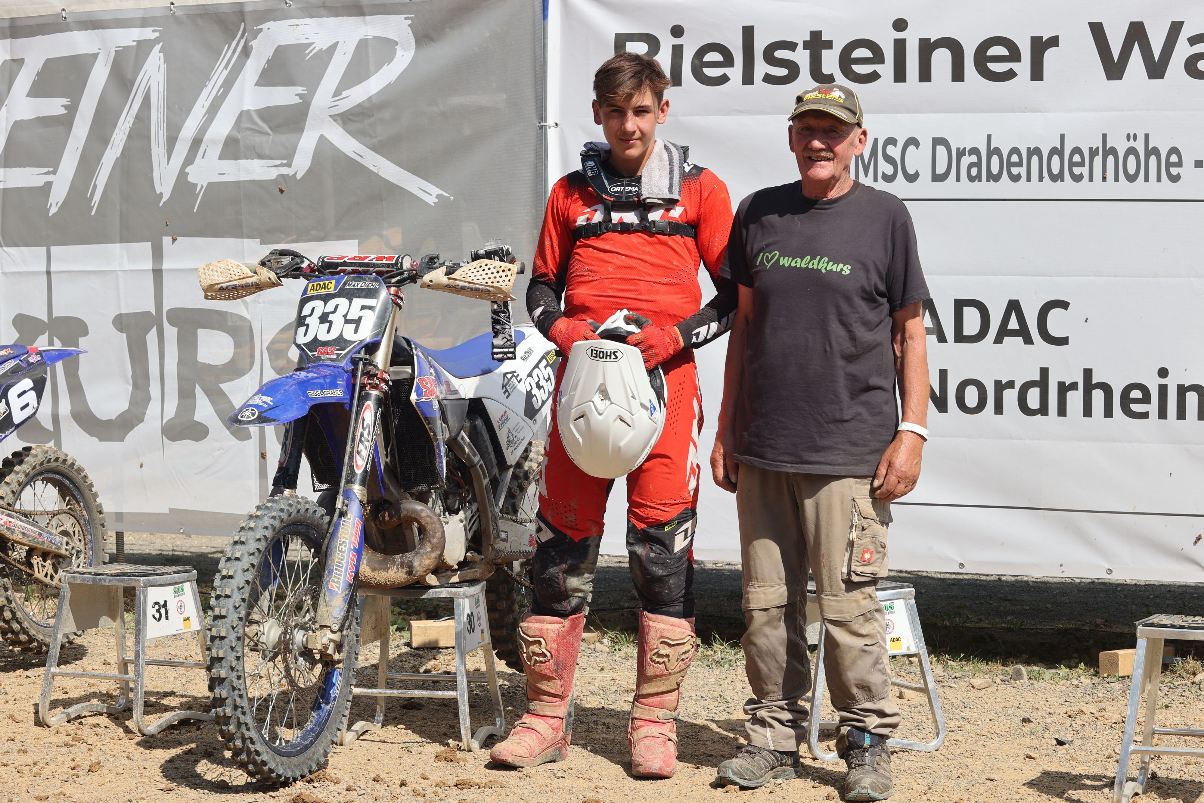 Motocrosser und Helfer stehen neben dem aufgebockten Motorrad.