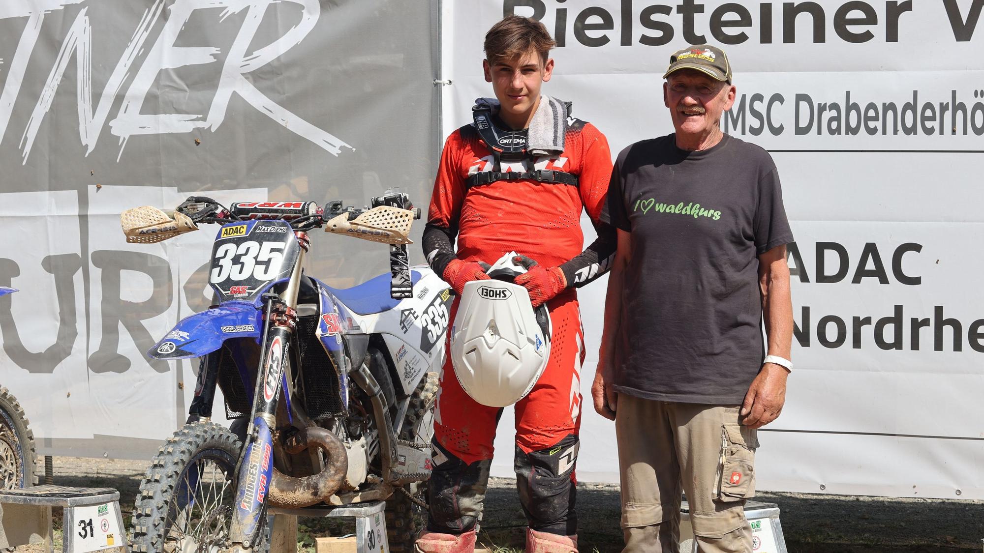 Motocrosser und Helfer stehen neben dem aufgebockten Motorrad.