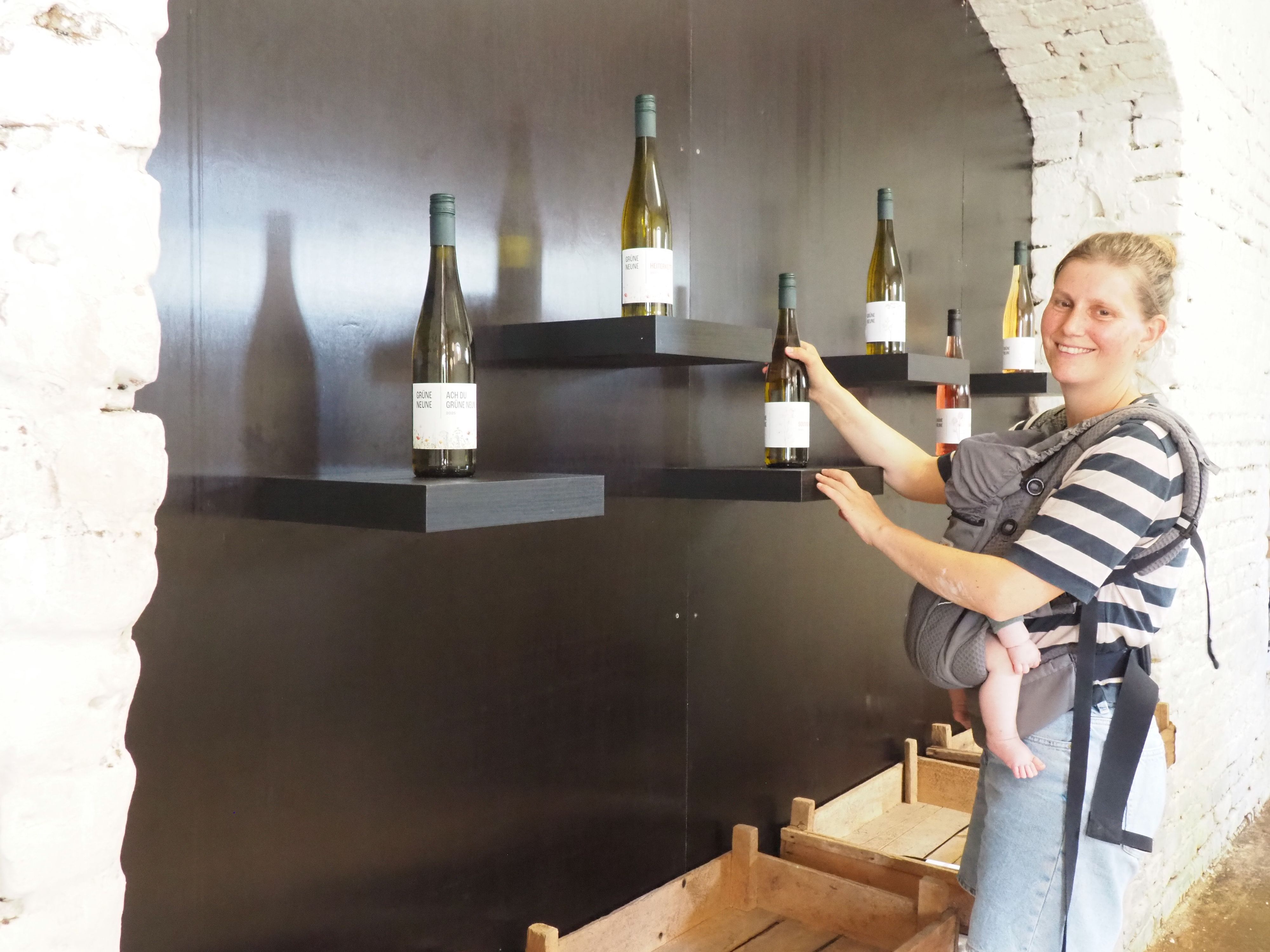 Eine Frau hält eine Flasche in einer Hand und steht vor einer Wand, an der einzelne Flaschen präsentiert werden.