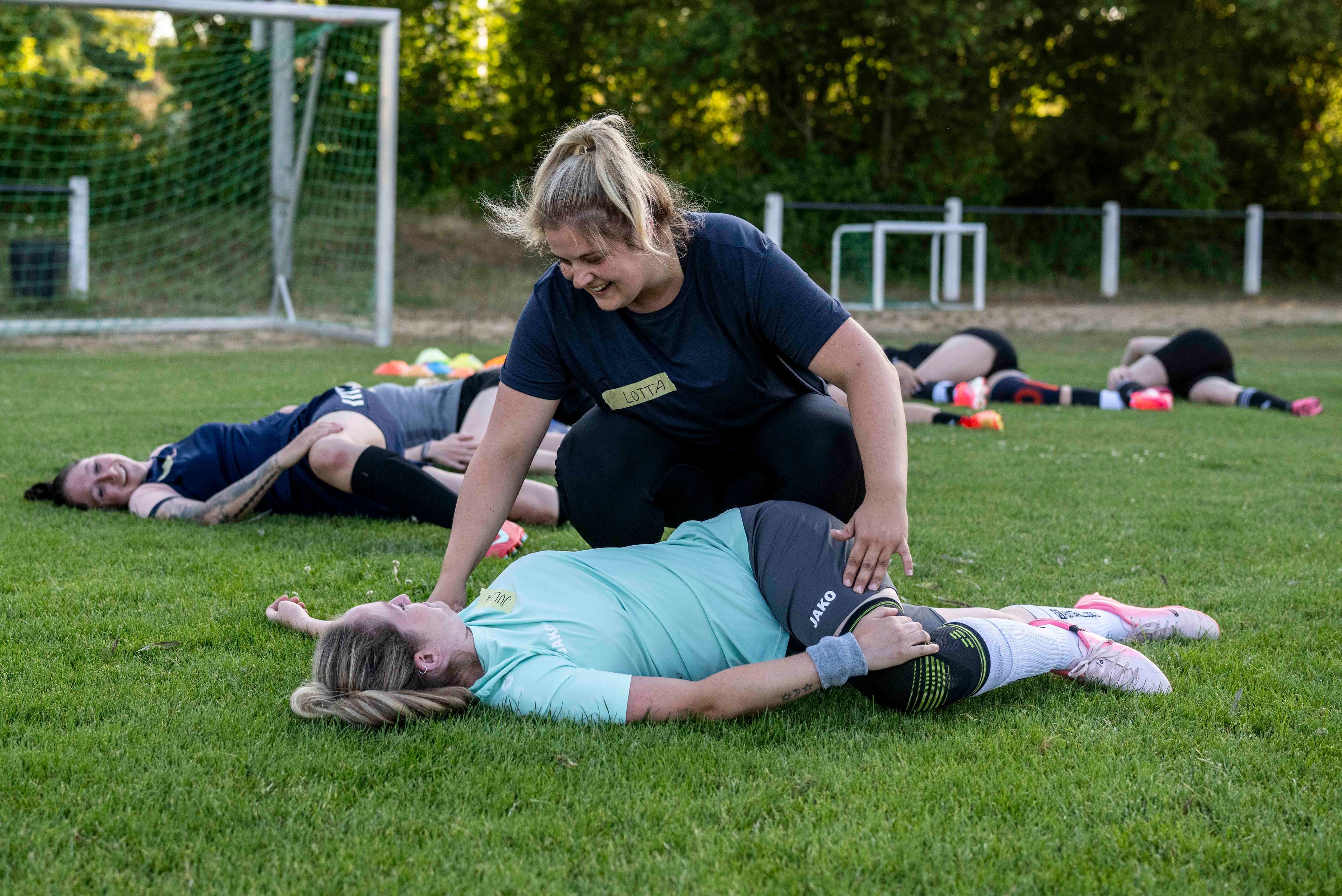 Schäfer hilft einer Spielerin, die auf dem Rasen liegt, bei einer Dehnübung. Im Hintergrund führen weitere Sportlerinnen diese Übung aus.