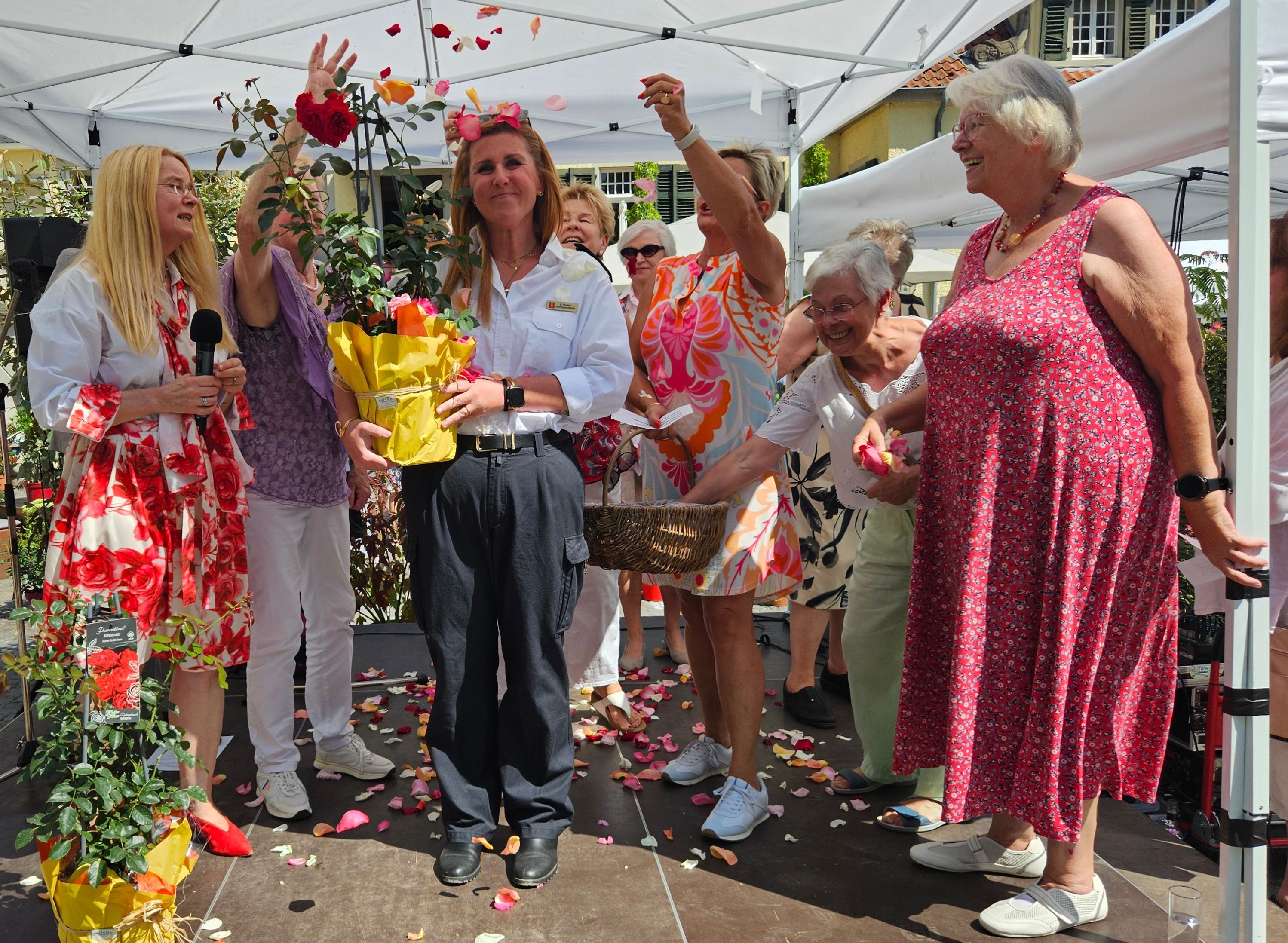 Eine Frau, mit einer Rose in der Hand, wird von anderen Frauen feierlich mit Rosenblütenblättern beworfen.
