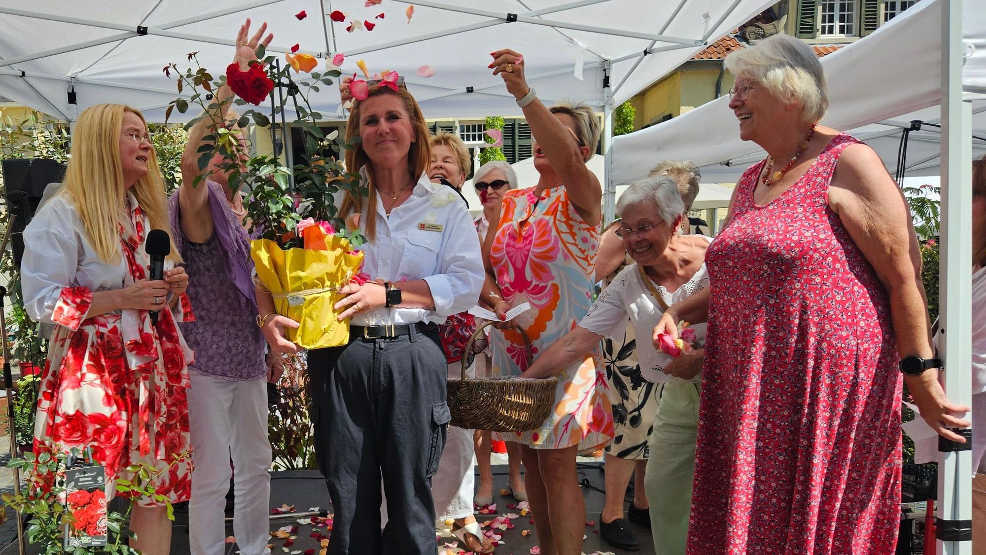 Eine Frau, mit einer Rose in der Hand, wird von anderen Frauen feierlich mit Rosenblütenblättern beworfen.