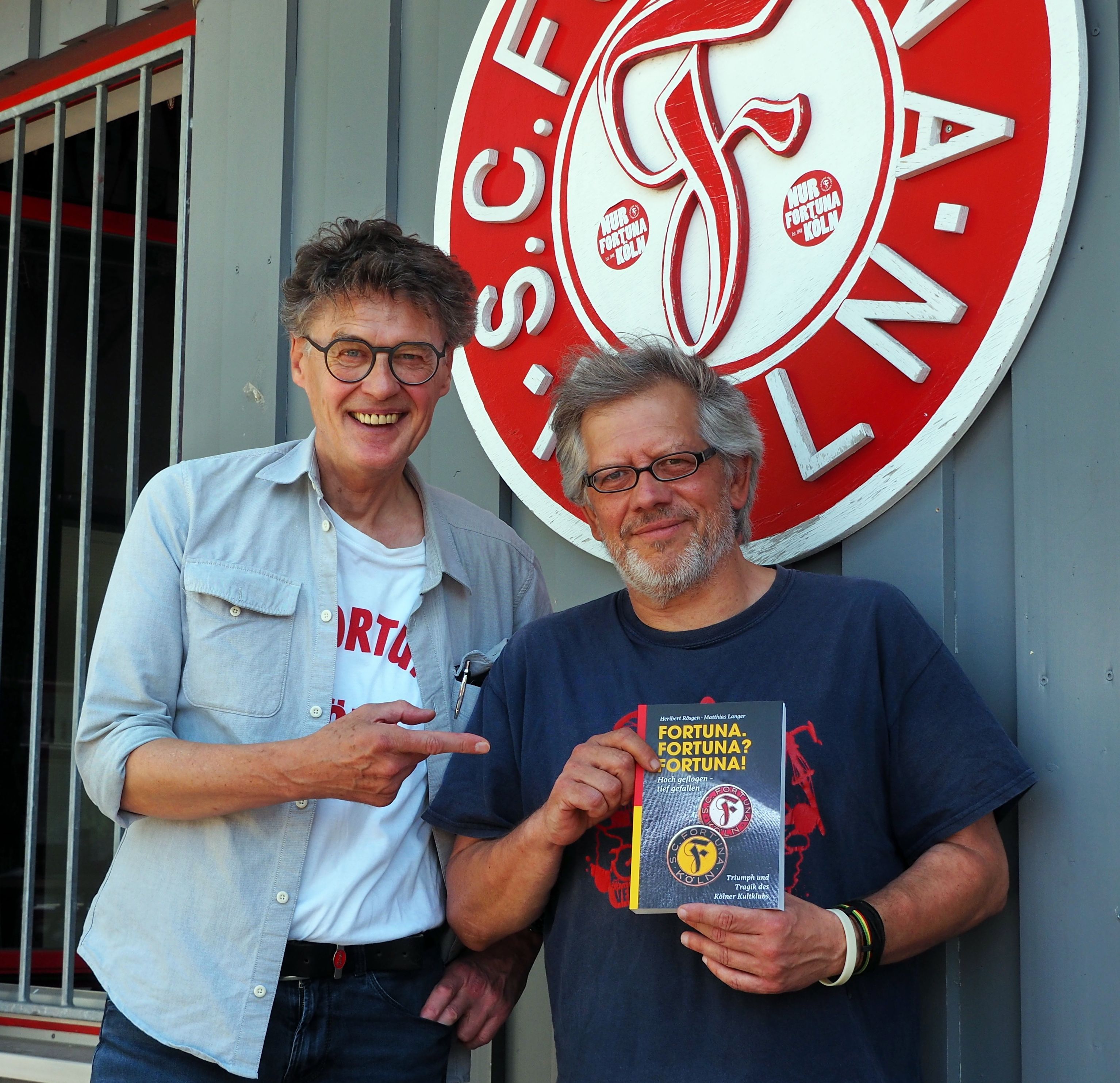 Zwei Männer stehen vor dem Vereinslogo von Fortuna Köln, der Mann rechts hat das neue Buch in der Hand.