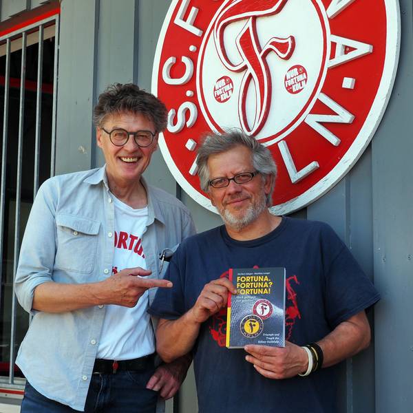 Zwei Männer stehen vor dem Vereinslogo von Fortuna Köln, der Mann rechts hat das neue Buch in der Hand.