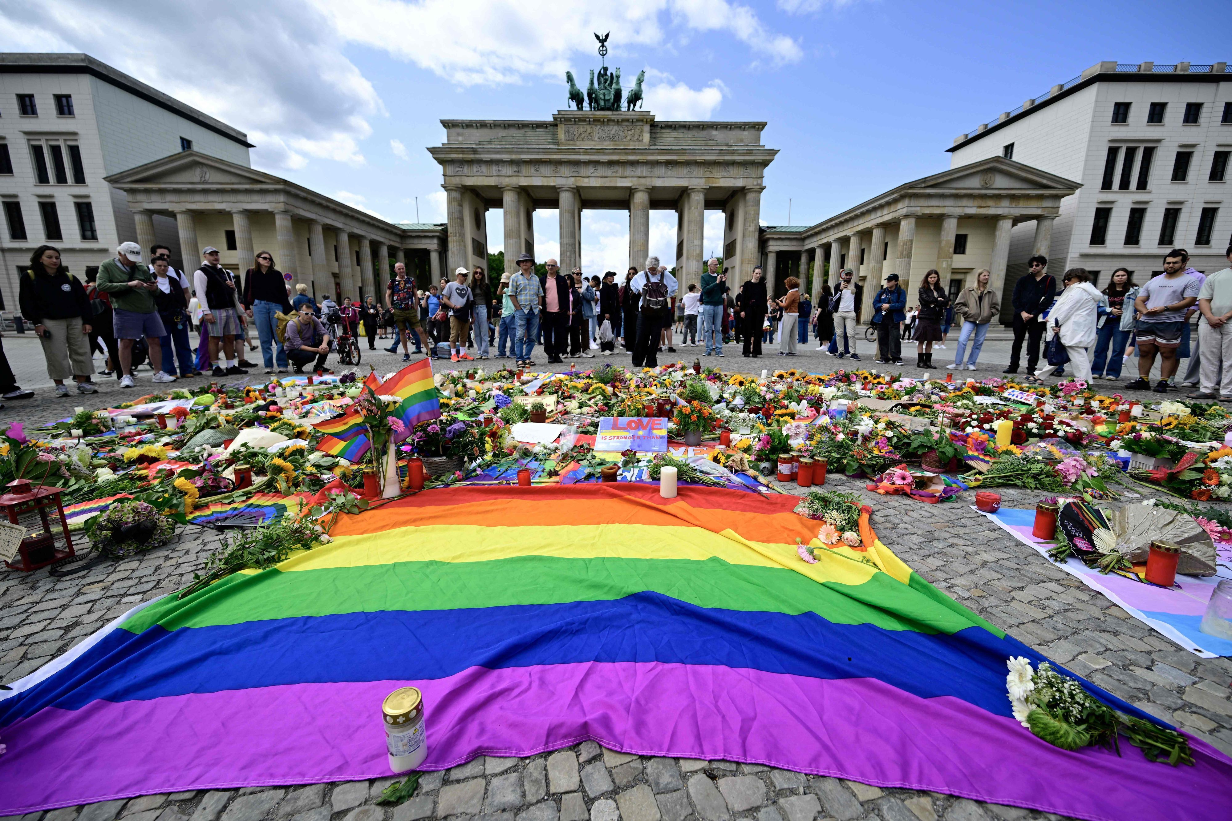 Trauernde nach dem CSD-Anchlag vom 25. Juli am Brandenburger Tor in Berlin. Im Vordergrund ist eine große Regenbogen-Flagge zu sehen, das Symbol der Queer-Bewegung