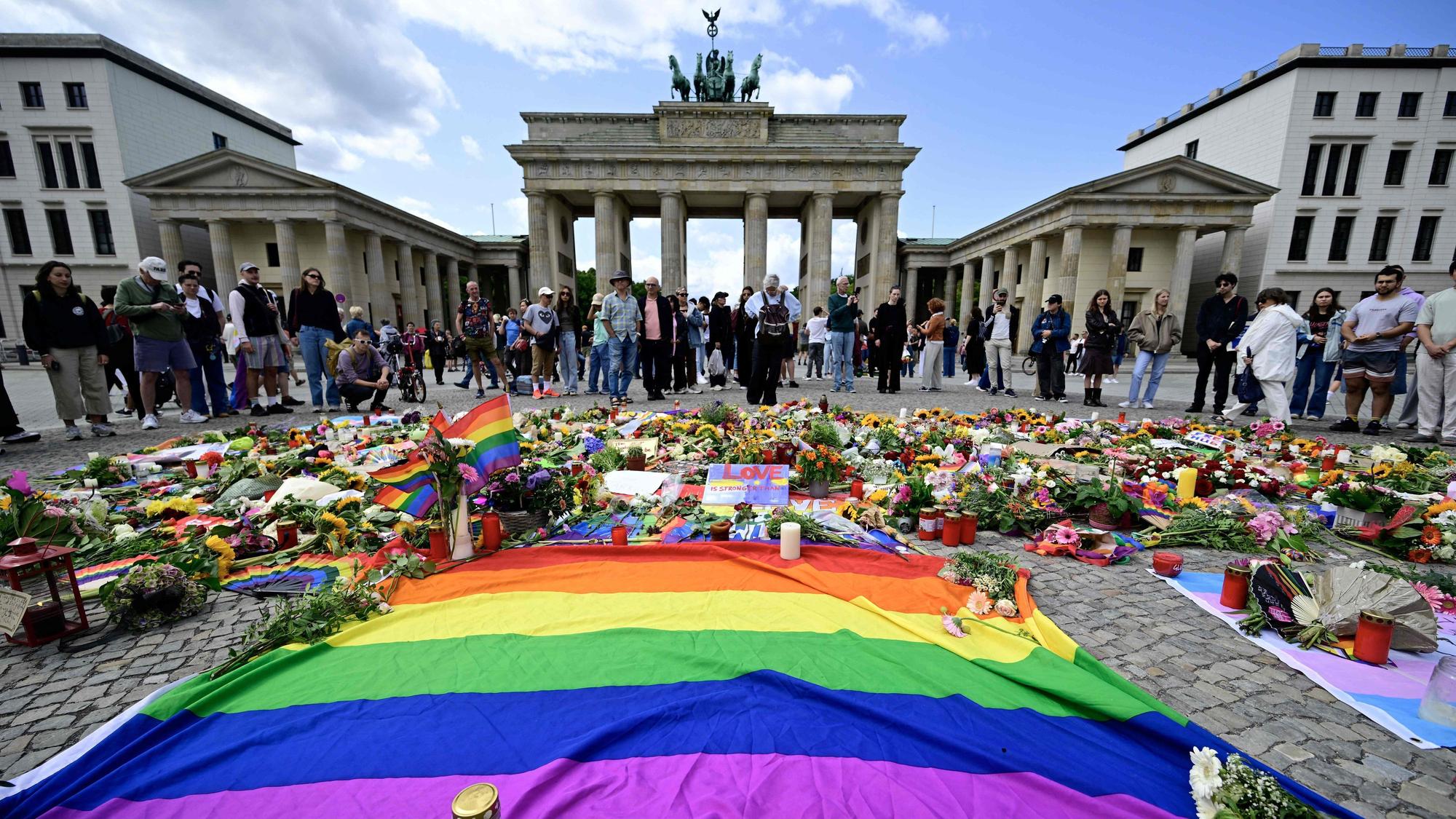 Trauernde nach dem CSD-Anchlag vom 25. Juli am Brandenburger Tor in Berlin. Im Vordergrund ist eine große Regenbogen-Flagge zu sehen, das Symbol der Queer-Bewegung