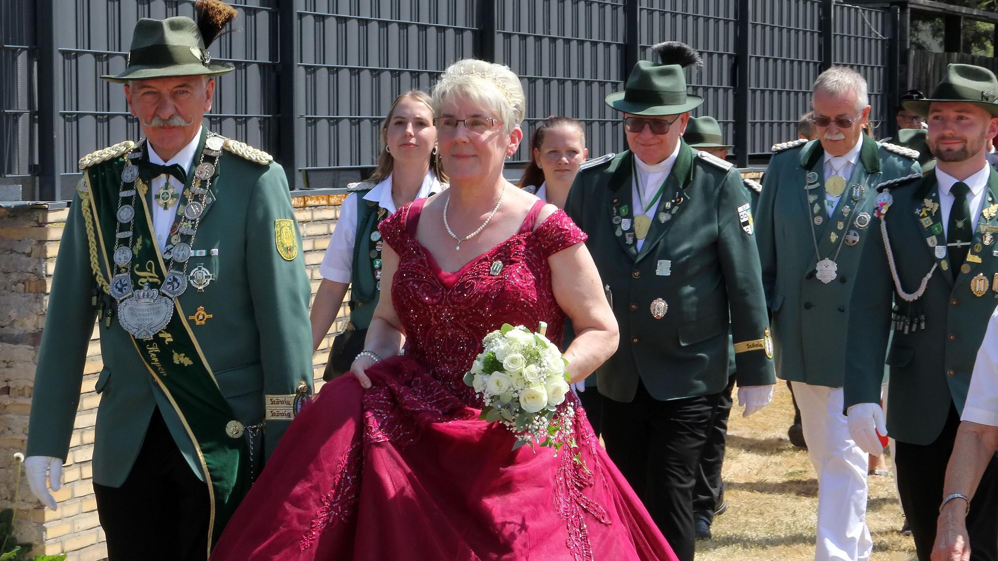 Männer und Frauen in Schützenuniformen und eine Frau in rotem Kleid gehen durch einen Ort.