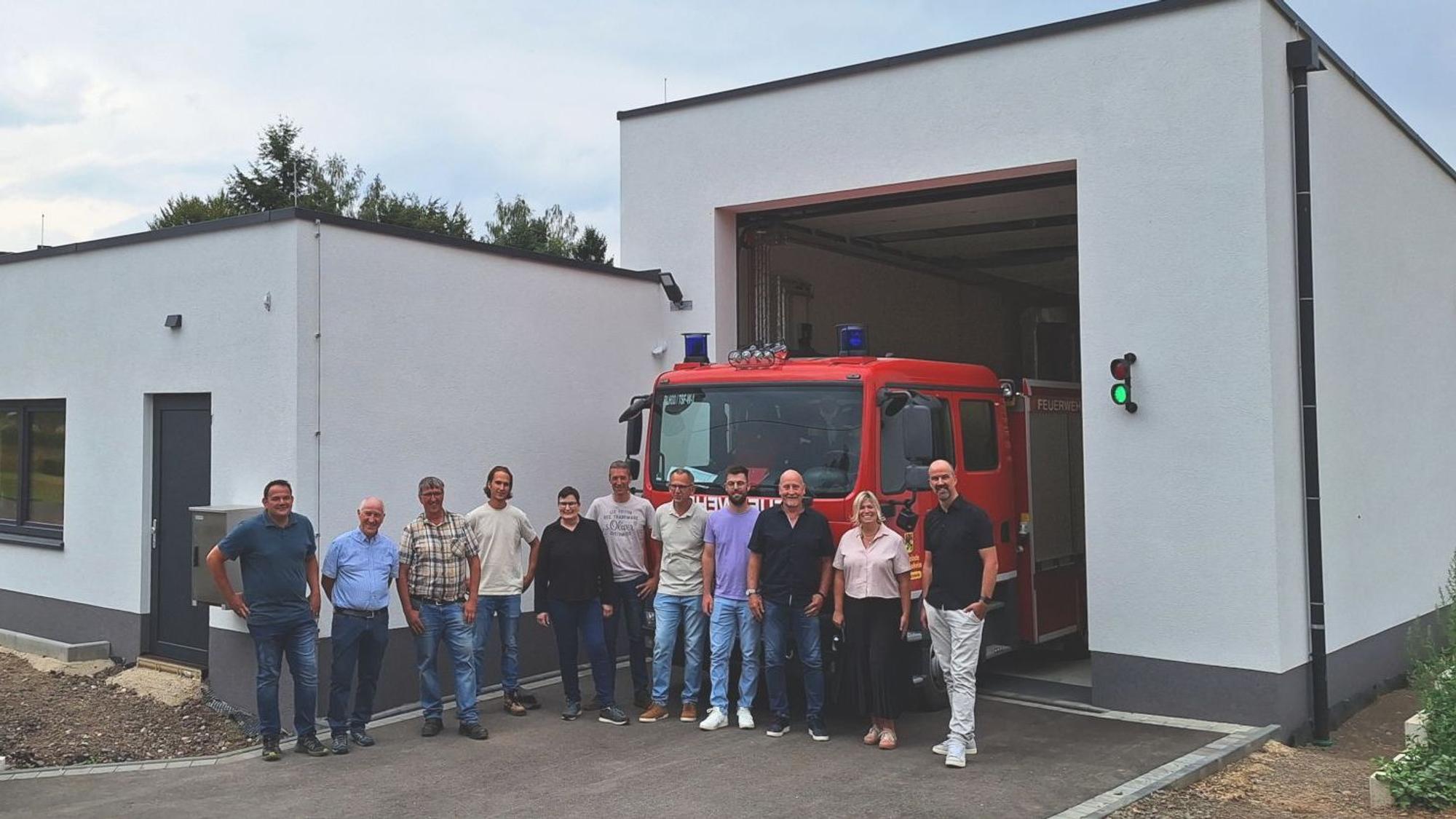 Die Genannten haben sich zum Gruppenbild vor der Wagenhalle mit dem Löschfahrzeug und dem Gerätehaus aufgestellt.