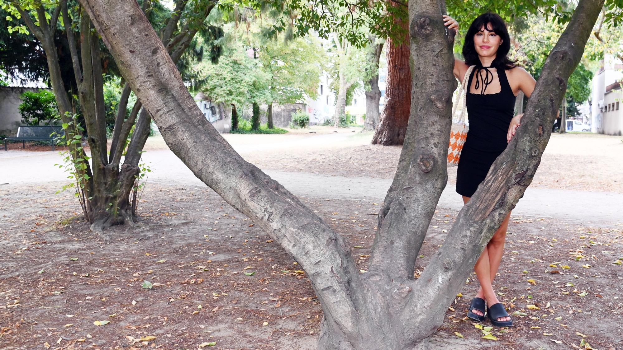 Eine Frau im schwarzen Kleid steht hinter einem Baum, der drei Stämme hat.