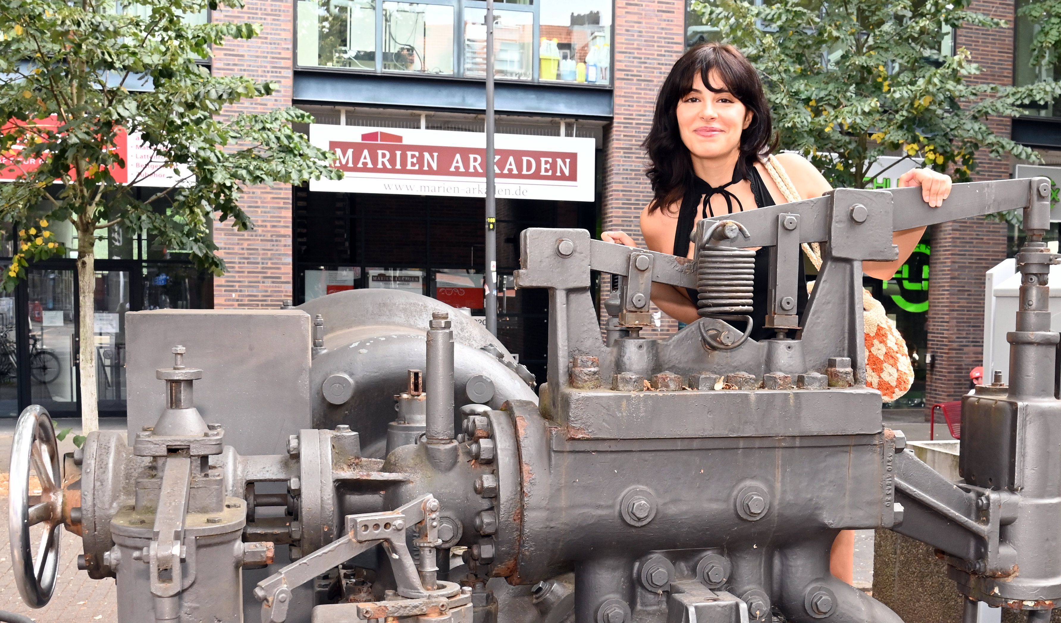 Eine junge Frau mit schwarzem Haar lugt über eine Industriemaschine, die als Denkmal an der Kalker Hauptstraße steht.
