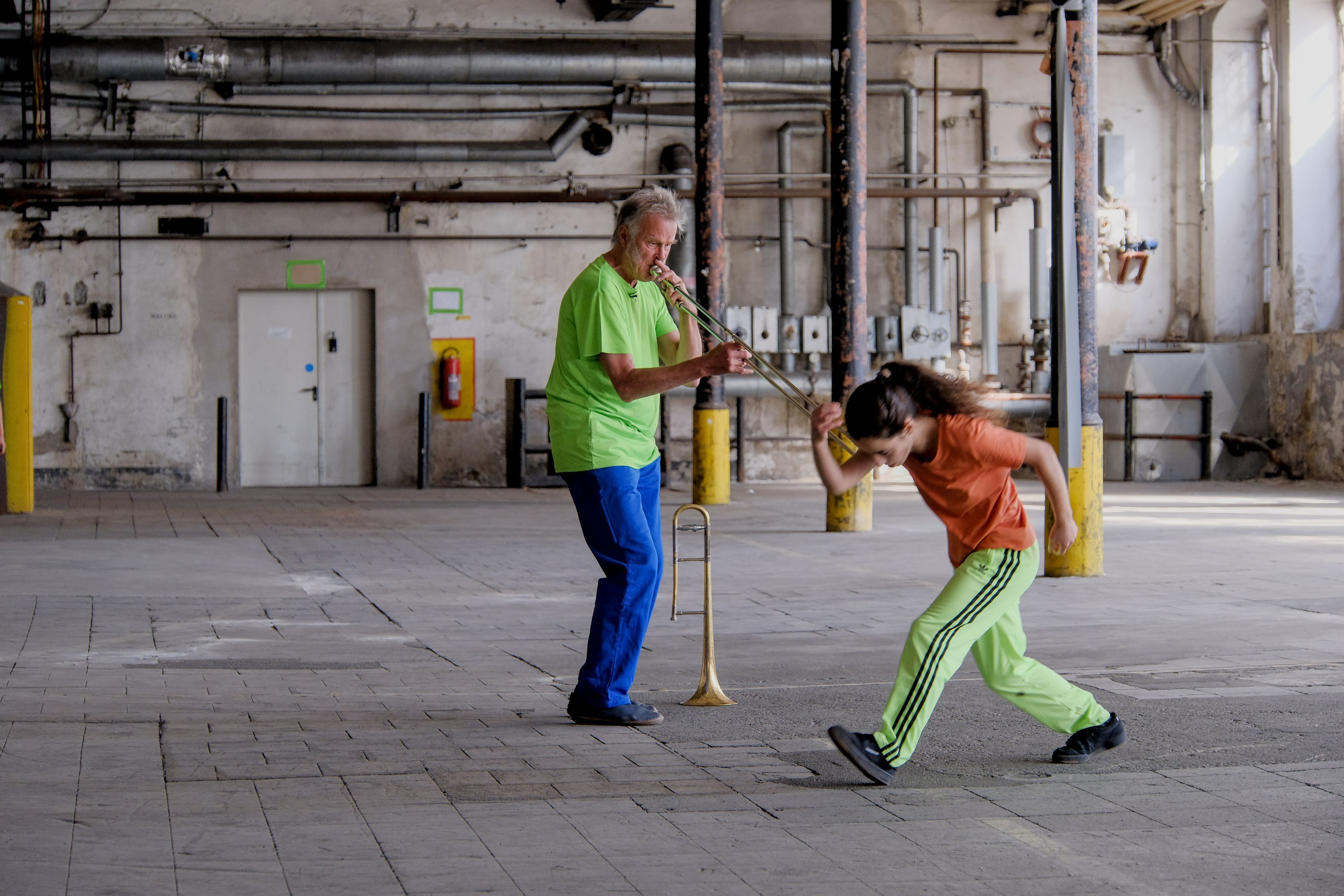 Ein Mann in neongrünem T-Shirt mit Posaune, eine Tänzerin in Orange und Neongrün macht einen großen Schritt