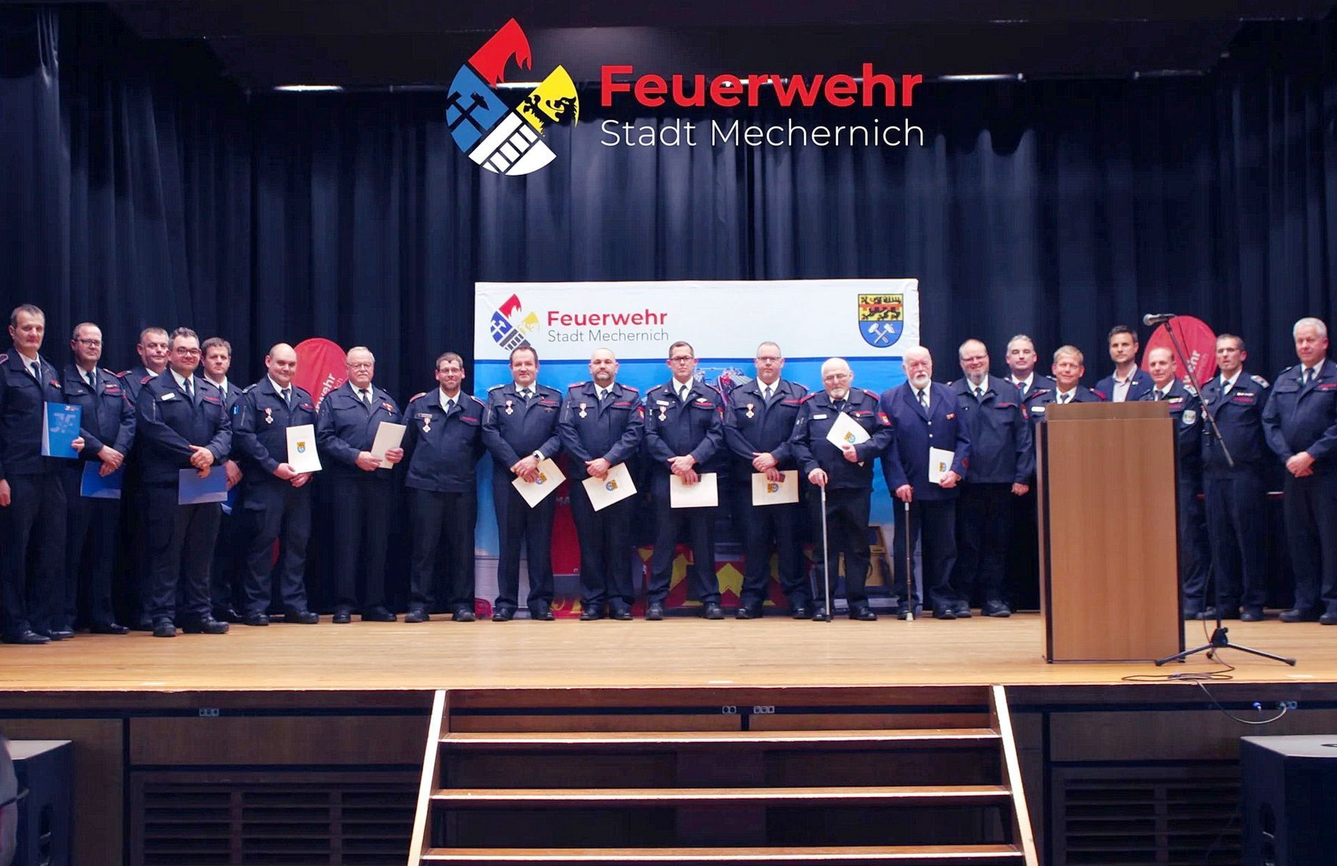 Zahlreiche Feuerwehrangehörige stehen in ihrer Ausgehuniform auf der Bühne der Mechernicher Schulaula. Einige halten Urkunden in der Hand.