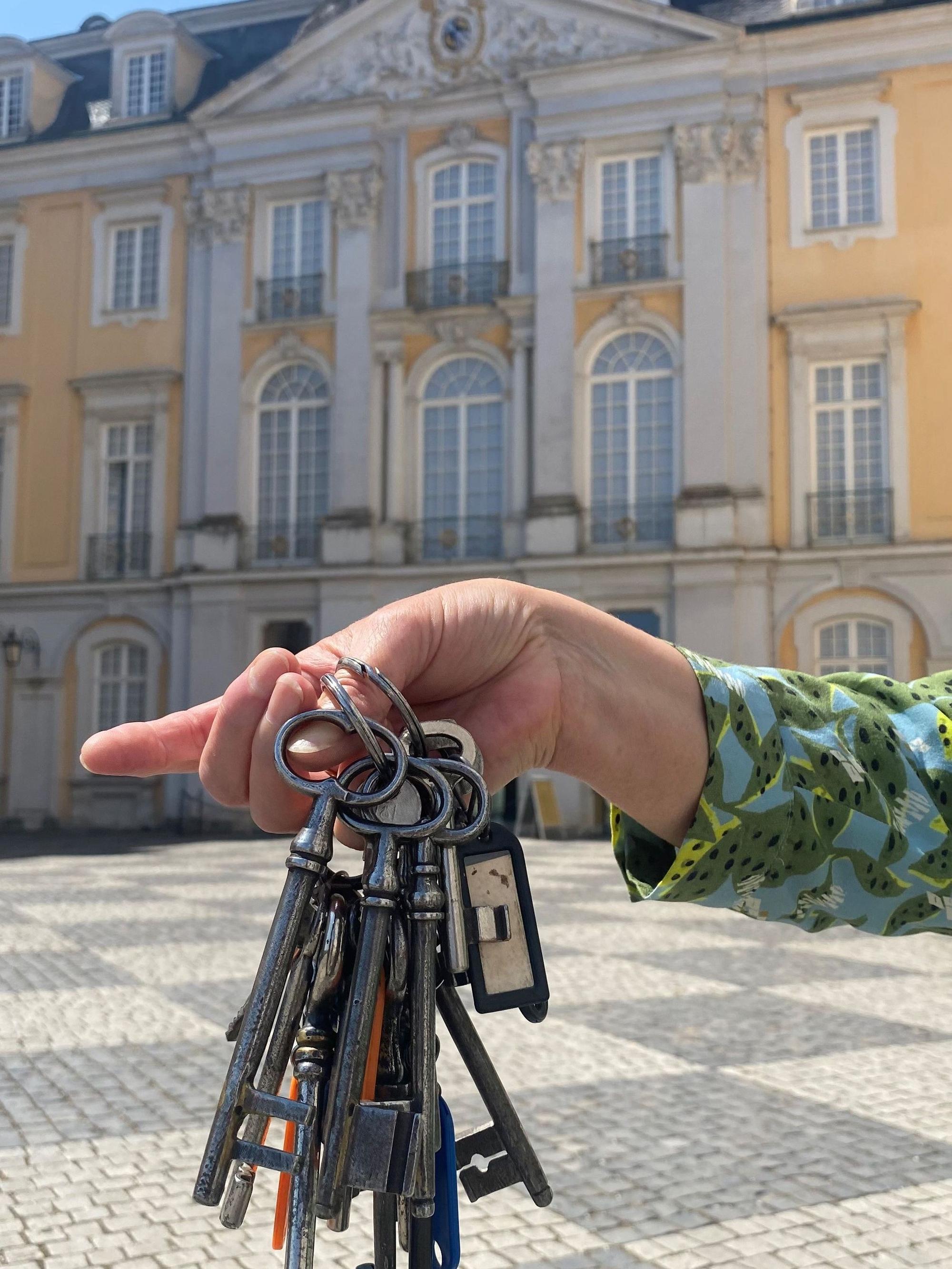 Zu sehen ist eine Hand, die ein Schlüsselbund hält. Im Hintergrund ist das Schloss Augustusburg zu sehen.