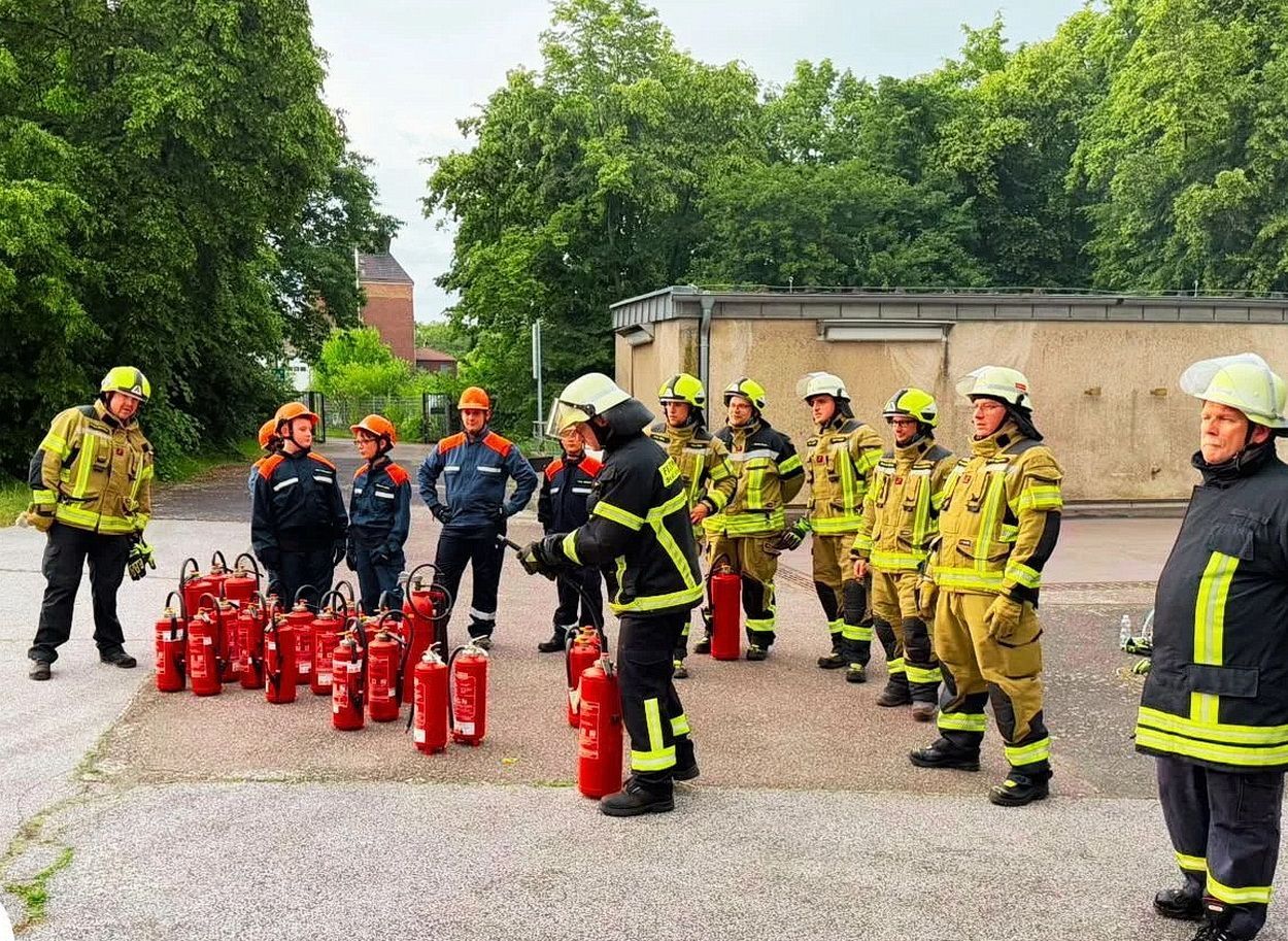 Feuerwehrleute und Mitglieder der Jugendfeuerwehr stehen in ihrer Einsatzkleidung hinter einer Batterie von Feuerlöschern.