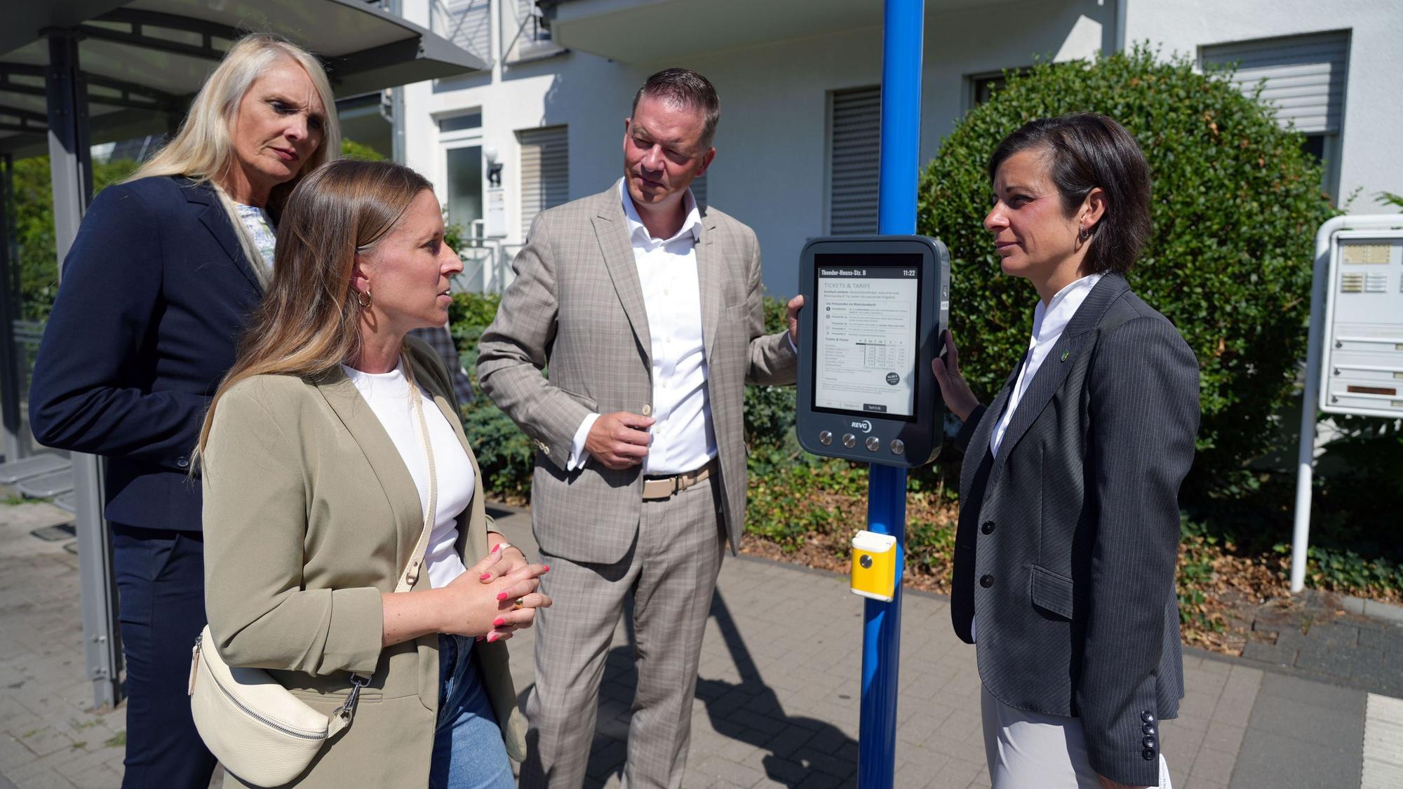 Eine Frau erklärt zwei Frauen und einem Mann eine digitale Anzeigetafel an einer Bushaltestelle.