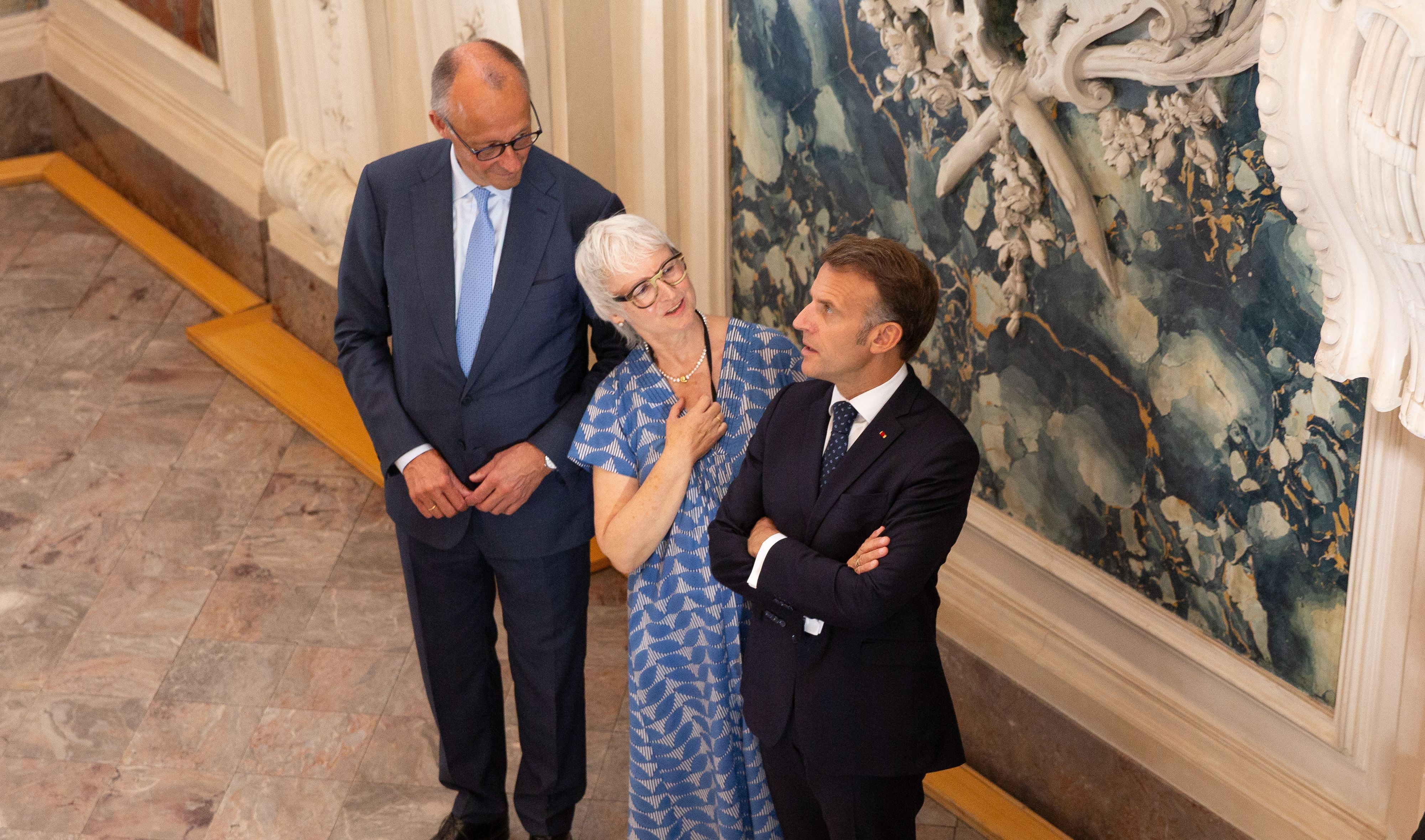 Zu sehen sind Regina Junga, Friedrich Merz und Emmanuel Macron im Treppenhaus des Schlosses.