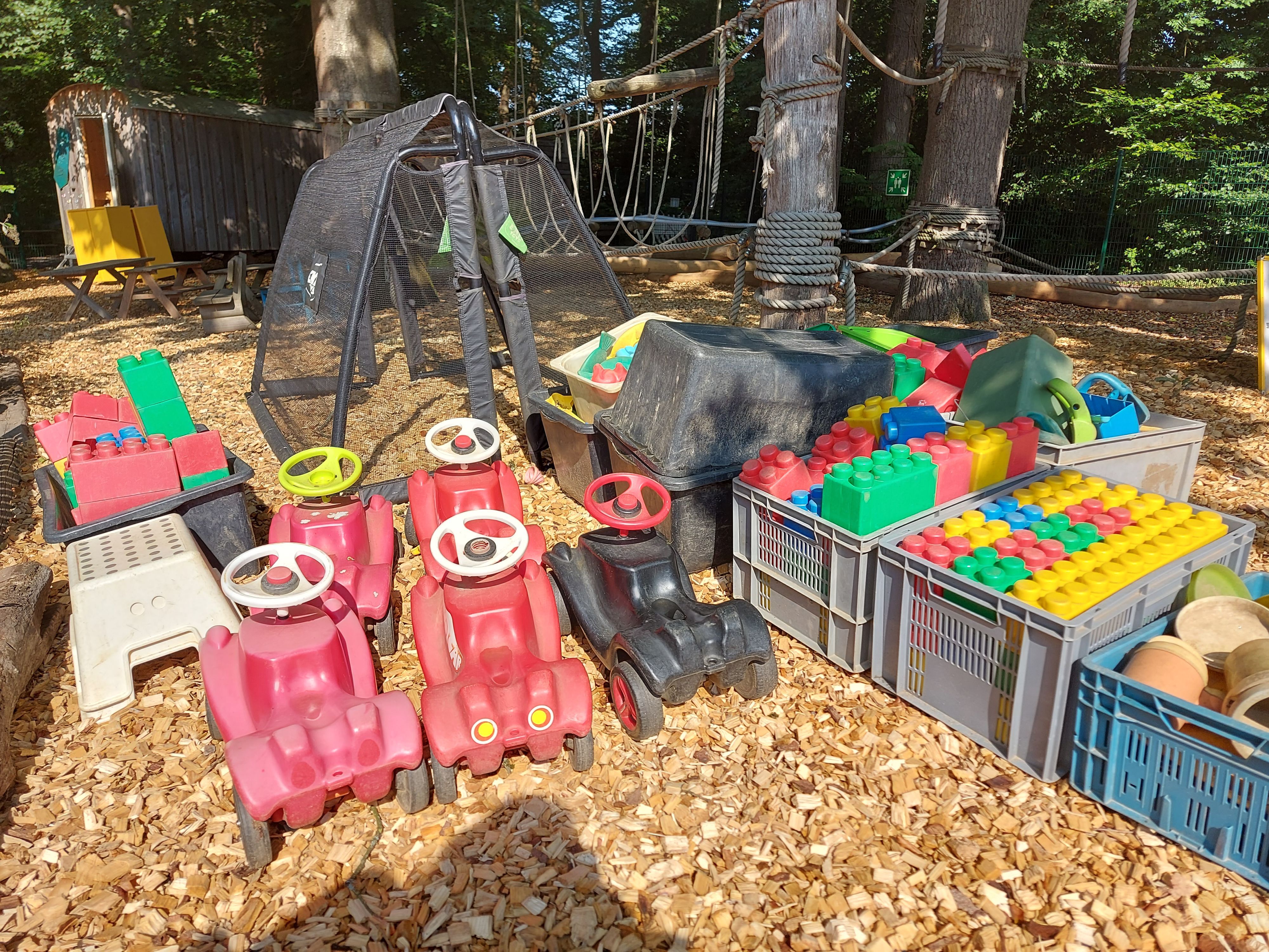 Fünf bunte Bobbycars und große Legosteine aus Kunststoff stehen auf groben Holzspänen.